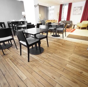 A modern dining area with wooden flooring, black chairs, and tables. There are potted plants and a red rug in the seating area. - Hotel Le Biarritz - Vichy (Vichy)