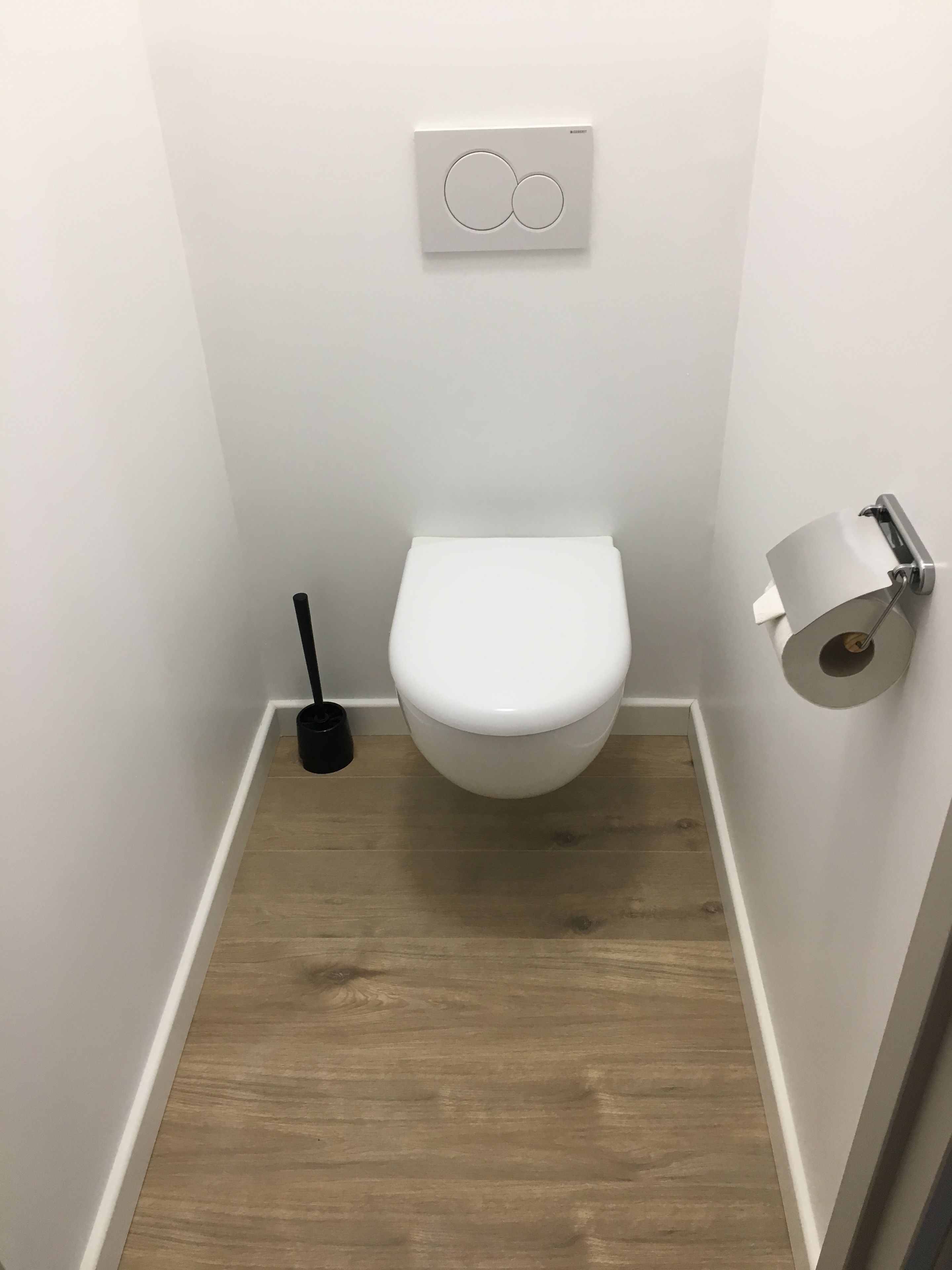 Un baño pequeño y moderno con inodoro blanco, dispensador de papel higiénico y un cepillo de limpieza negro.
