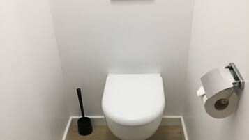 Un baño pequeño y moderno con inodoro blanco, dispensador de papel higiénico y un cepillo de limpieza negro.