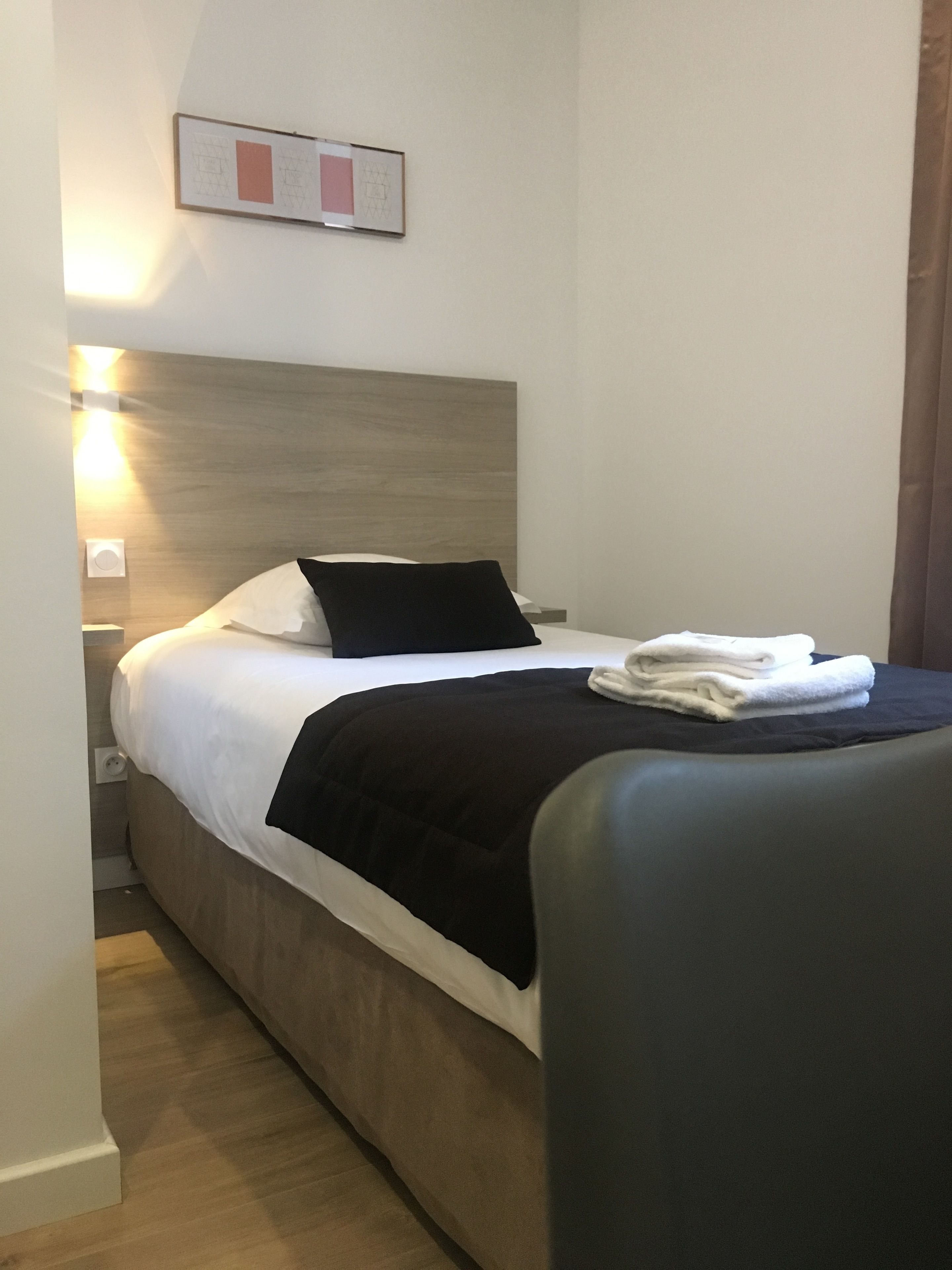 Un lit bien fait, avec du linge de lit blanc et noir, une serviette pliée et un tableau encadré accroché au mur.
