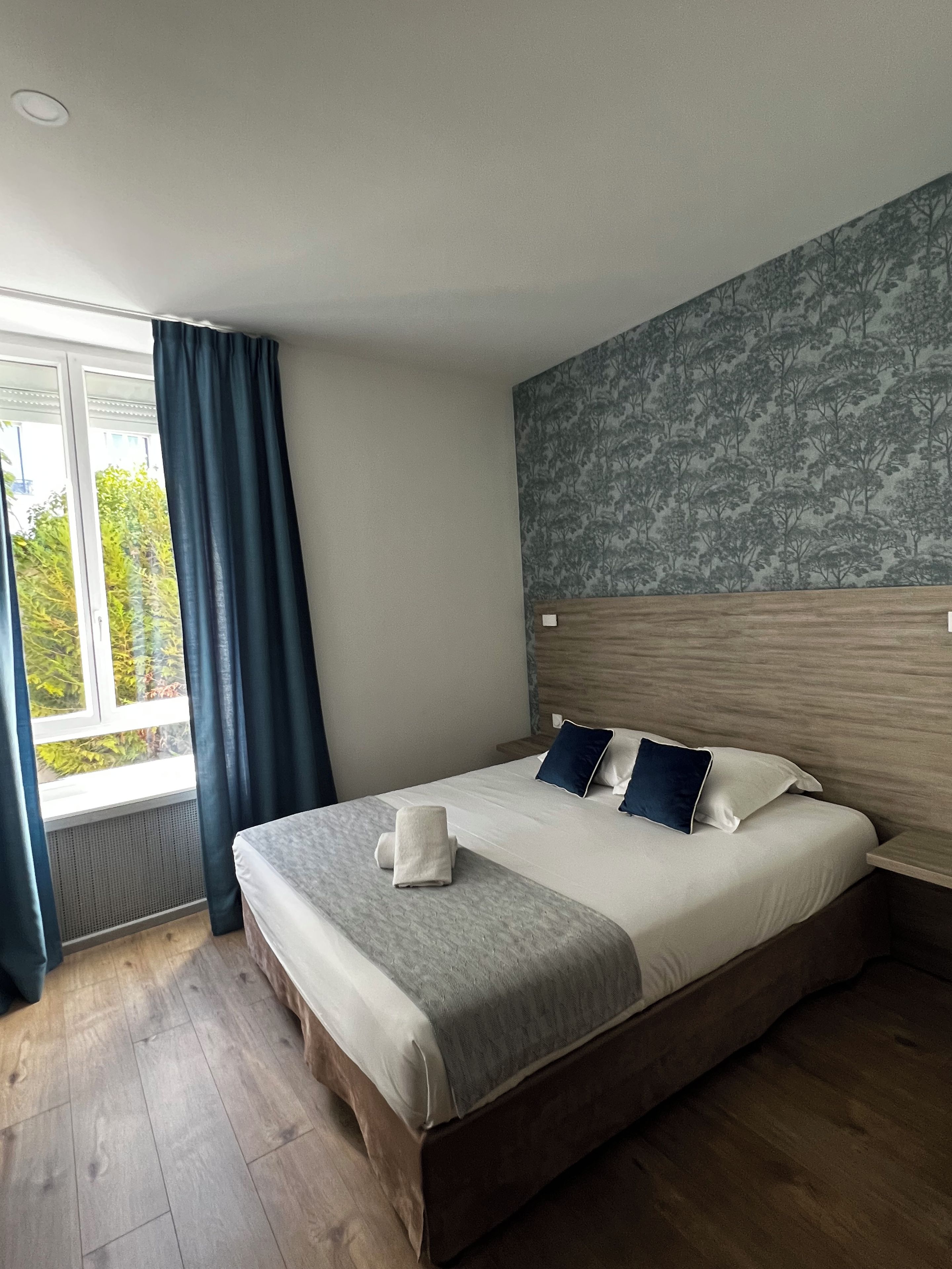 A hotel room with a large bed, a window with curtains, a patterned wall, and a wooden floor.
