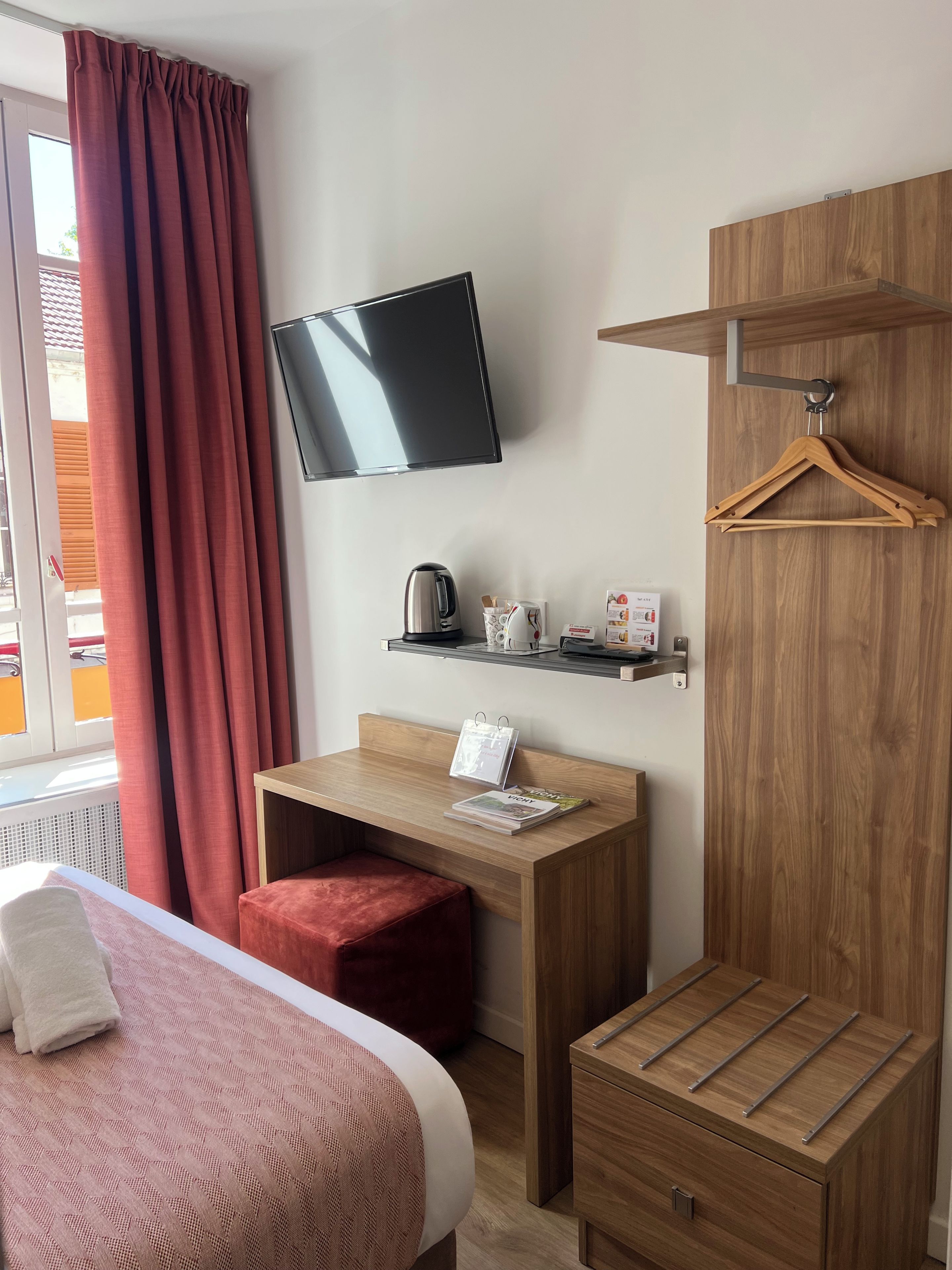 A hotel room with a bed, a desk with a chair, a television mounted on the wall, a wooden wardrobe, and a window with curtains.