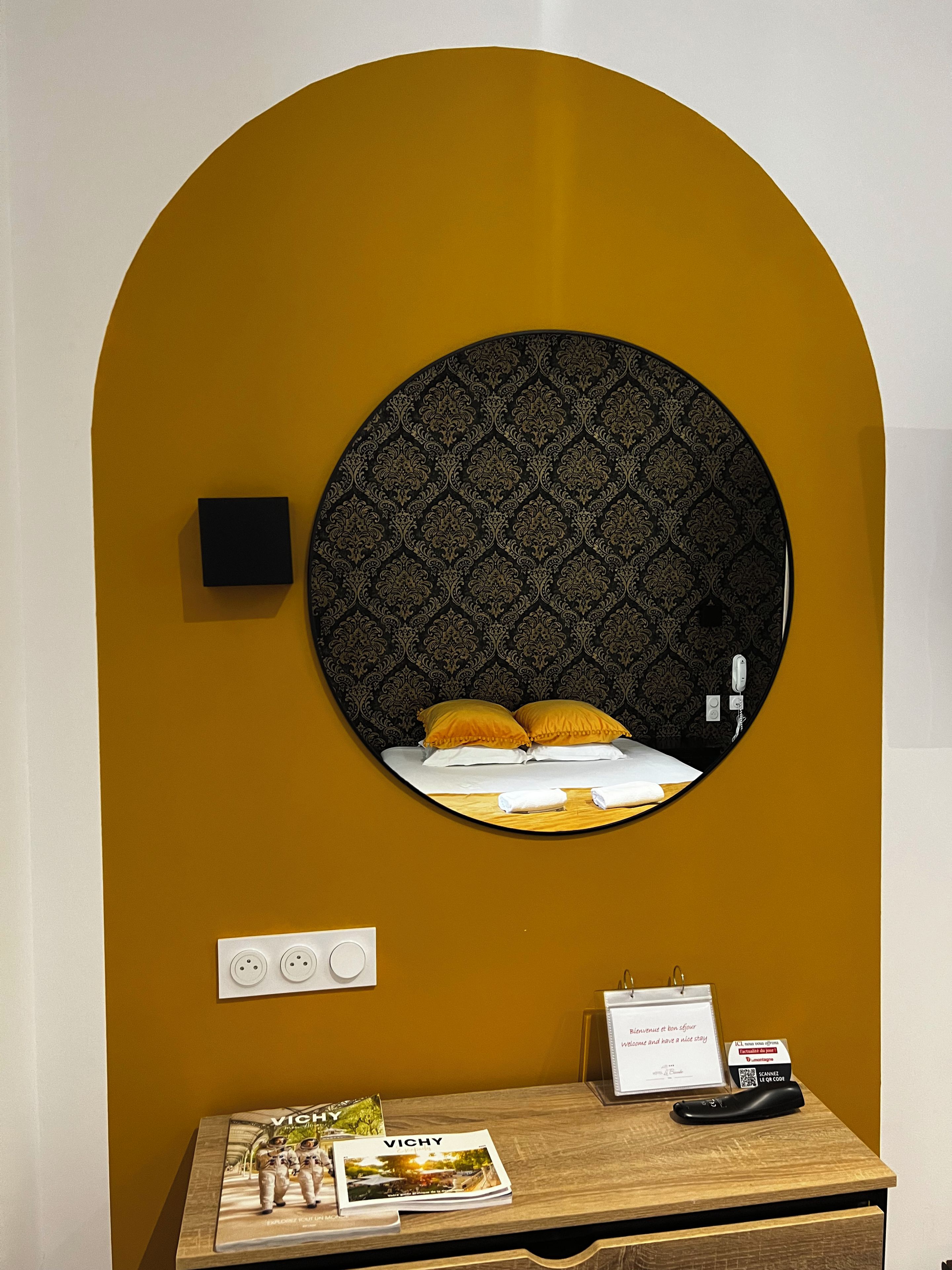 A hotel room with a circular mirror, a bed with yellow pillows, a wooden desk with a magazine, and a sign with a QR code.