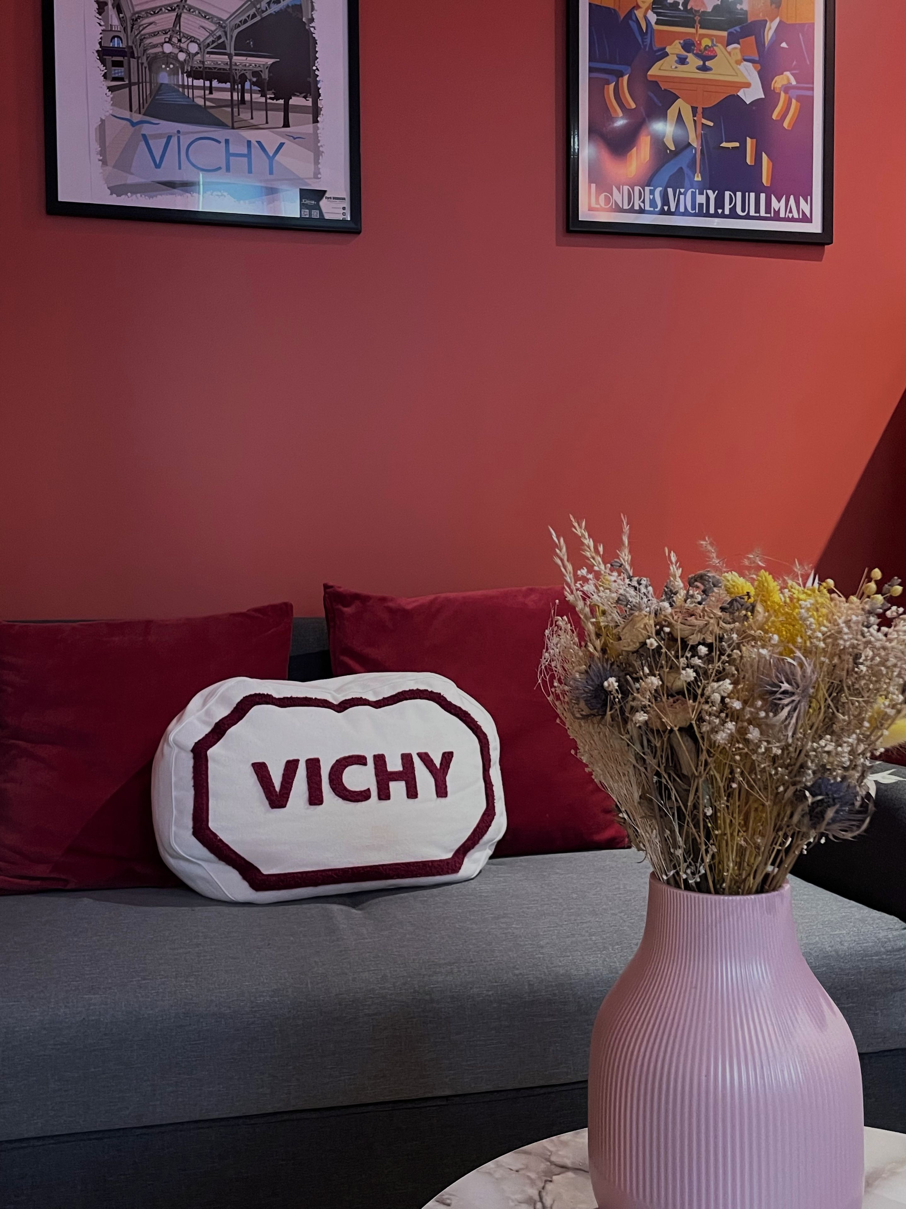 A room with a red wall, a grey sofa, a white pillow with 'VICHY' embroidered, a pink vase with dried flowers, and two framed artworks.