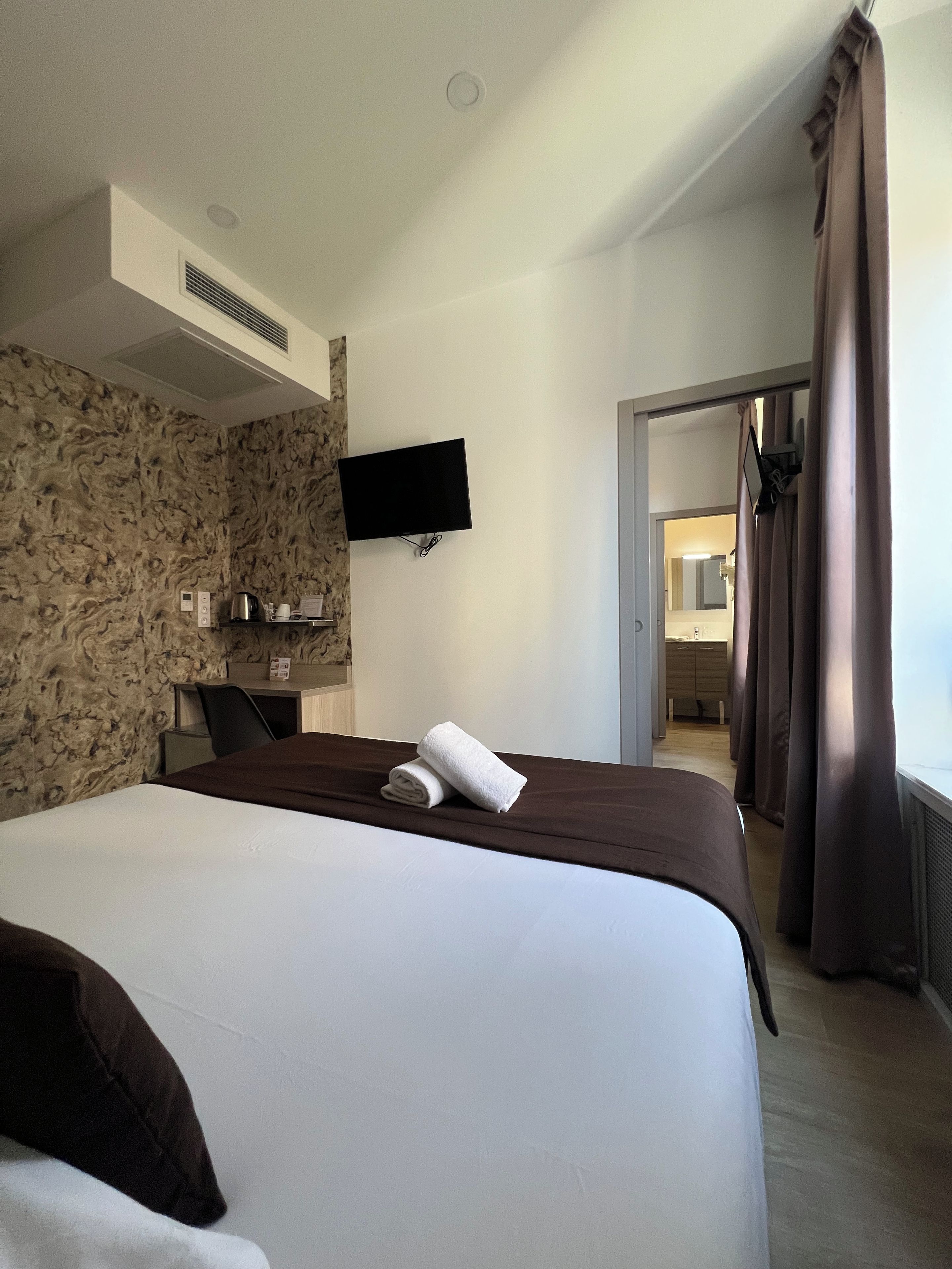 Ein Hotelzimmer mit einem Bett, einem flachbildfernseher an der Wand, einem Schreibtisch mit Stuhl und einem durch eine offene Tür einsehbaren Badezimmer.