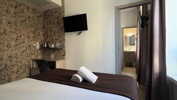 Ein Hotelzimmer mit einem Bett, einem flachbildfernseher an der Wand, einem Schreibtisch mit Stuhl und einem durch eine offene Tür einsehbaren Badezimmer.