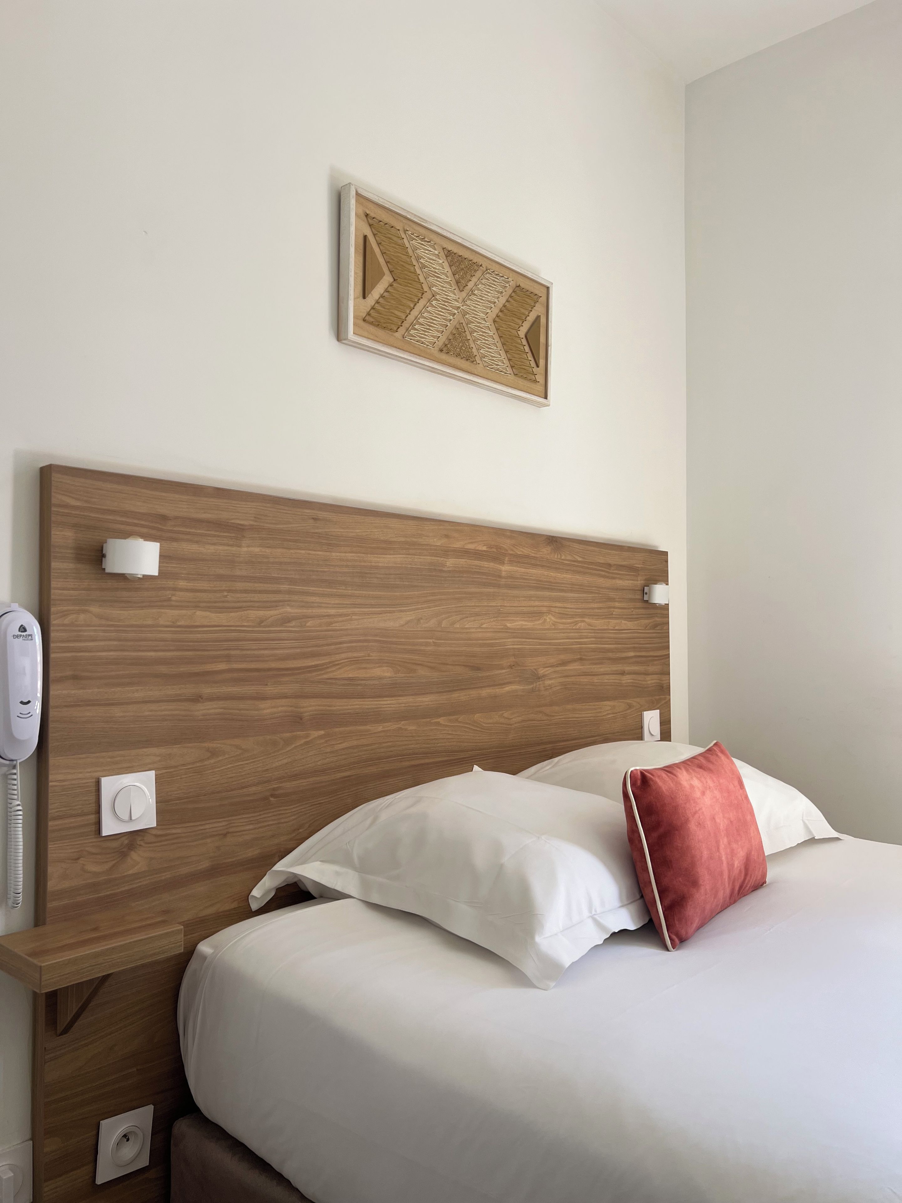 Une chambre d’hôtel avec une tête de lit en bois, un téléphone sur la table de chevet, une prise électrique et un lit avec une literie blanche et un oreiller rouge.