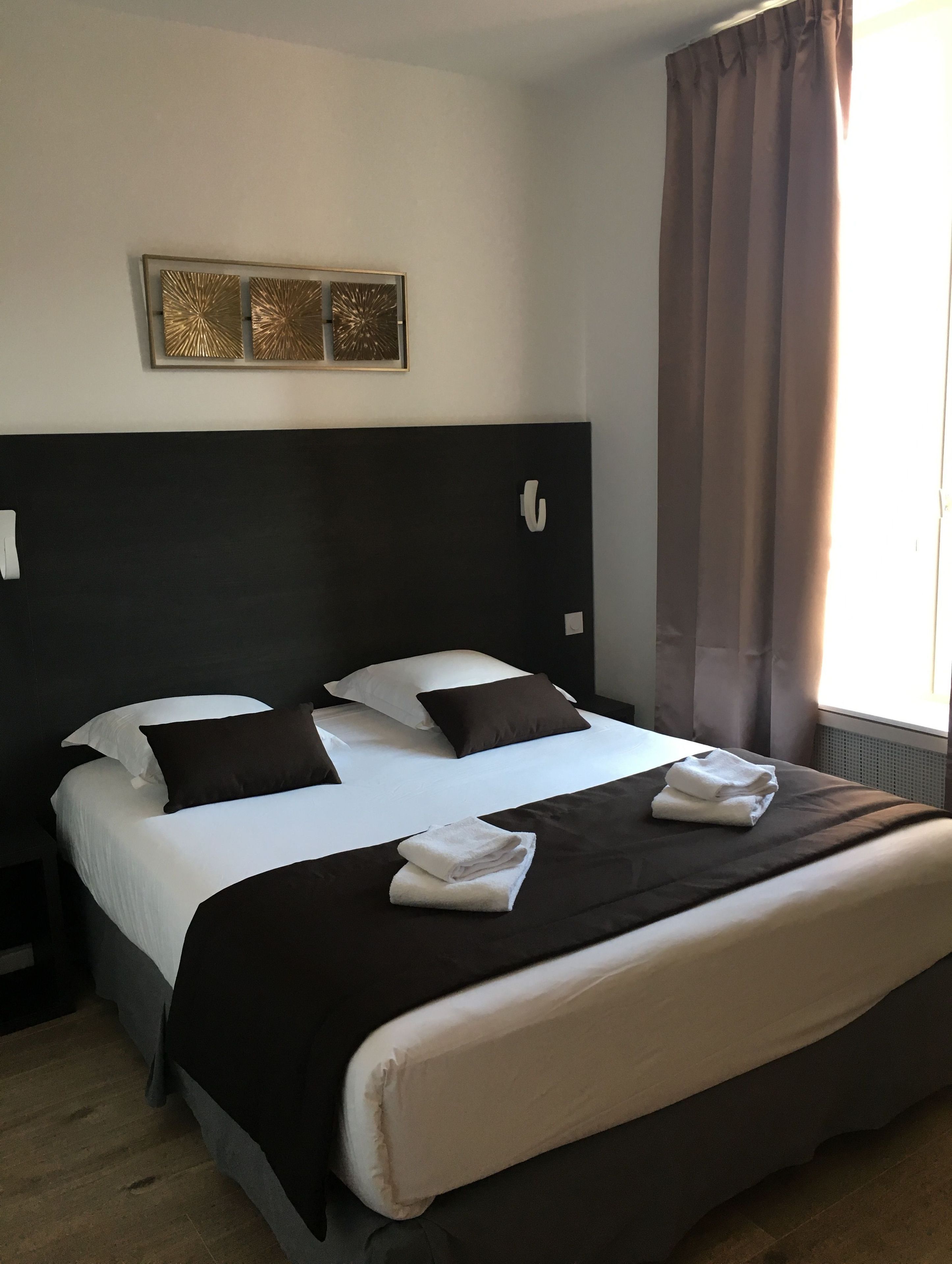 A hotel room with a large bed, two pillows, a headboard, a bedside table, and a window with curtains.