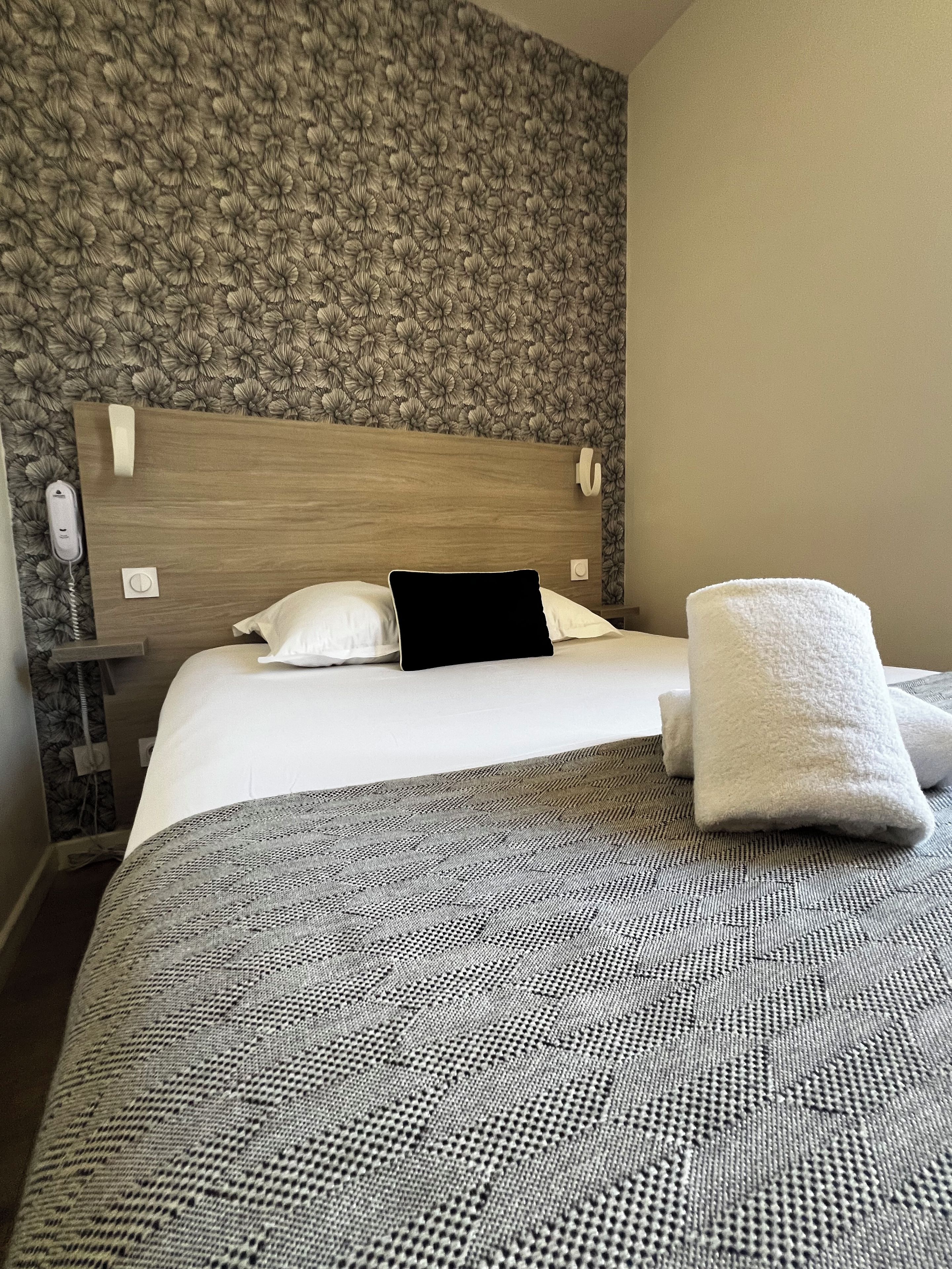 Une chambre d’hôtel avec un papier peint à motifs, une tête de lit en bois, un lit avec du linge de maison blanc, un oreiller noir et une serviette blanche.