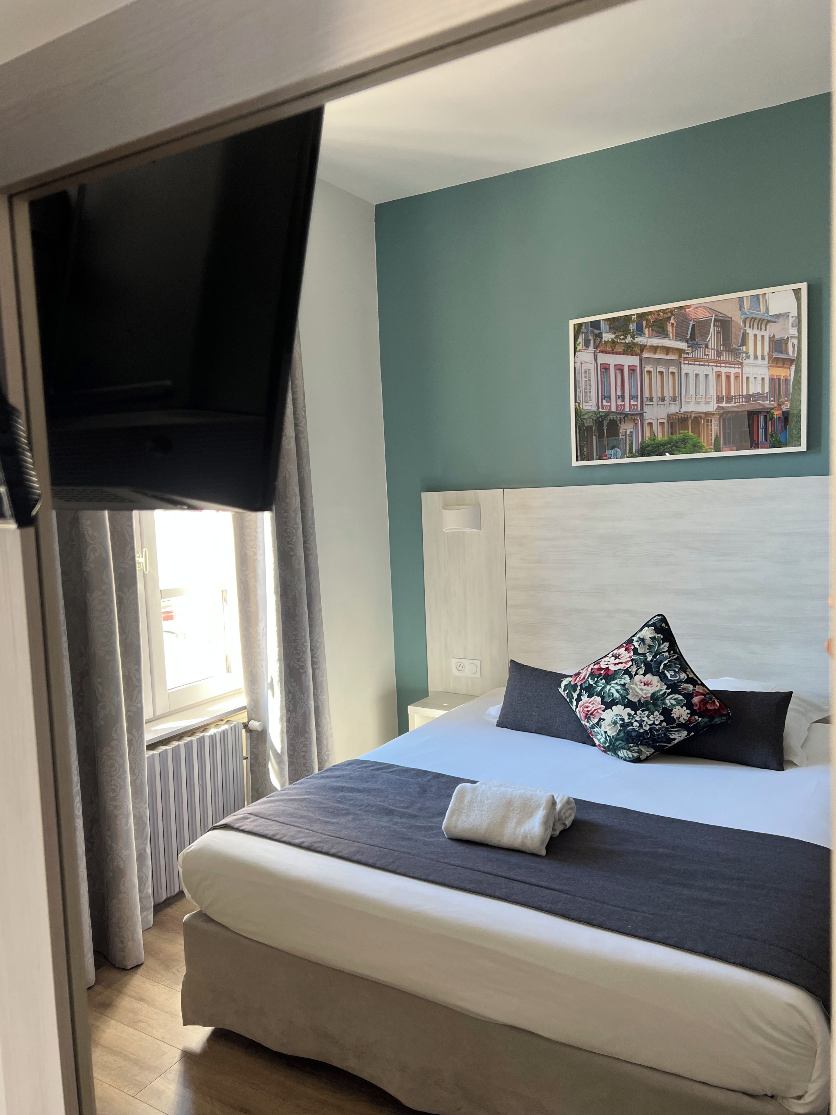 Une chambre d’hôtel avec un lit, un téléviseur à écran plat fixé au mur, une fenêtre avec des rideaux et un tableau au-dessus du lit.