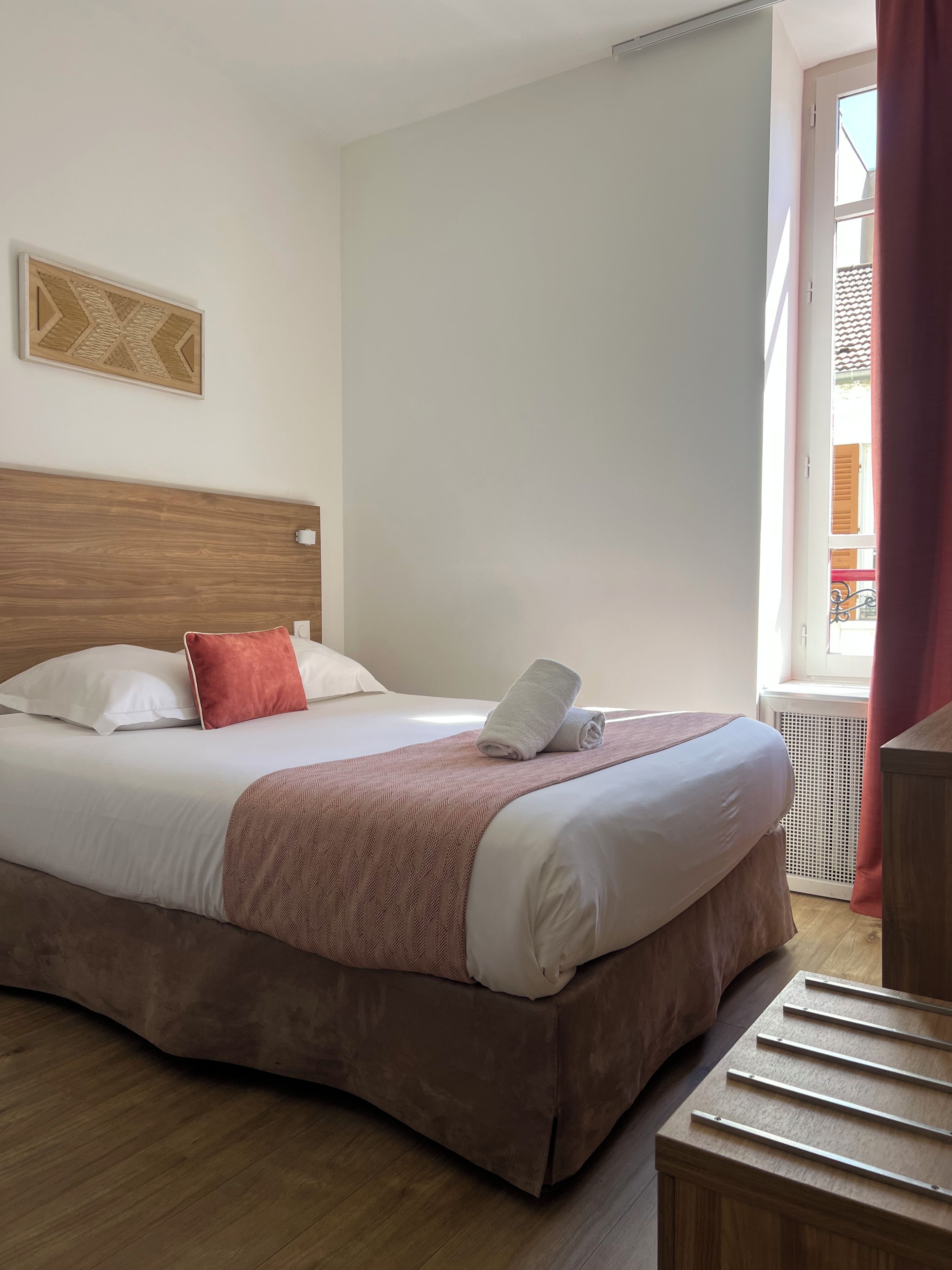 A hotel room with a bed, a headboard, a window with curtains, and a wooden floor.