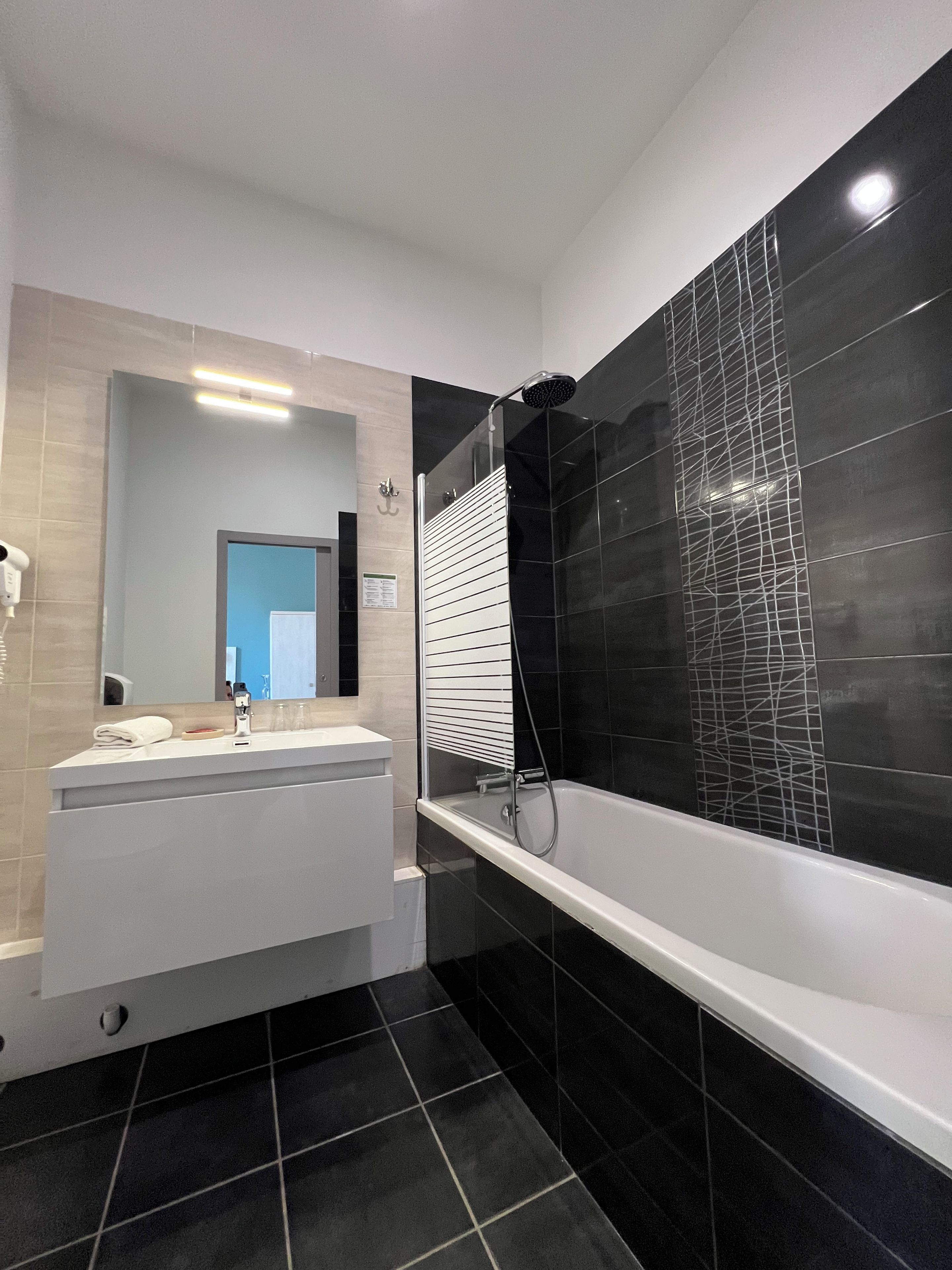 Un baño moderno con tocador blanco, un espejo grande, un box de ducha con azulejos negros y piso con diseño de azulejos negros.