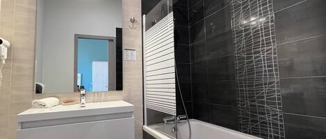 Un baño moderno con tocador blanco, un espejo grande, un box de ducha con azulejos negros y piso con diseño de azulejos negros.