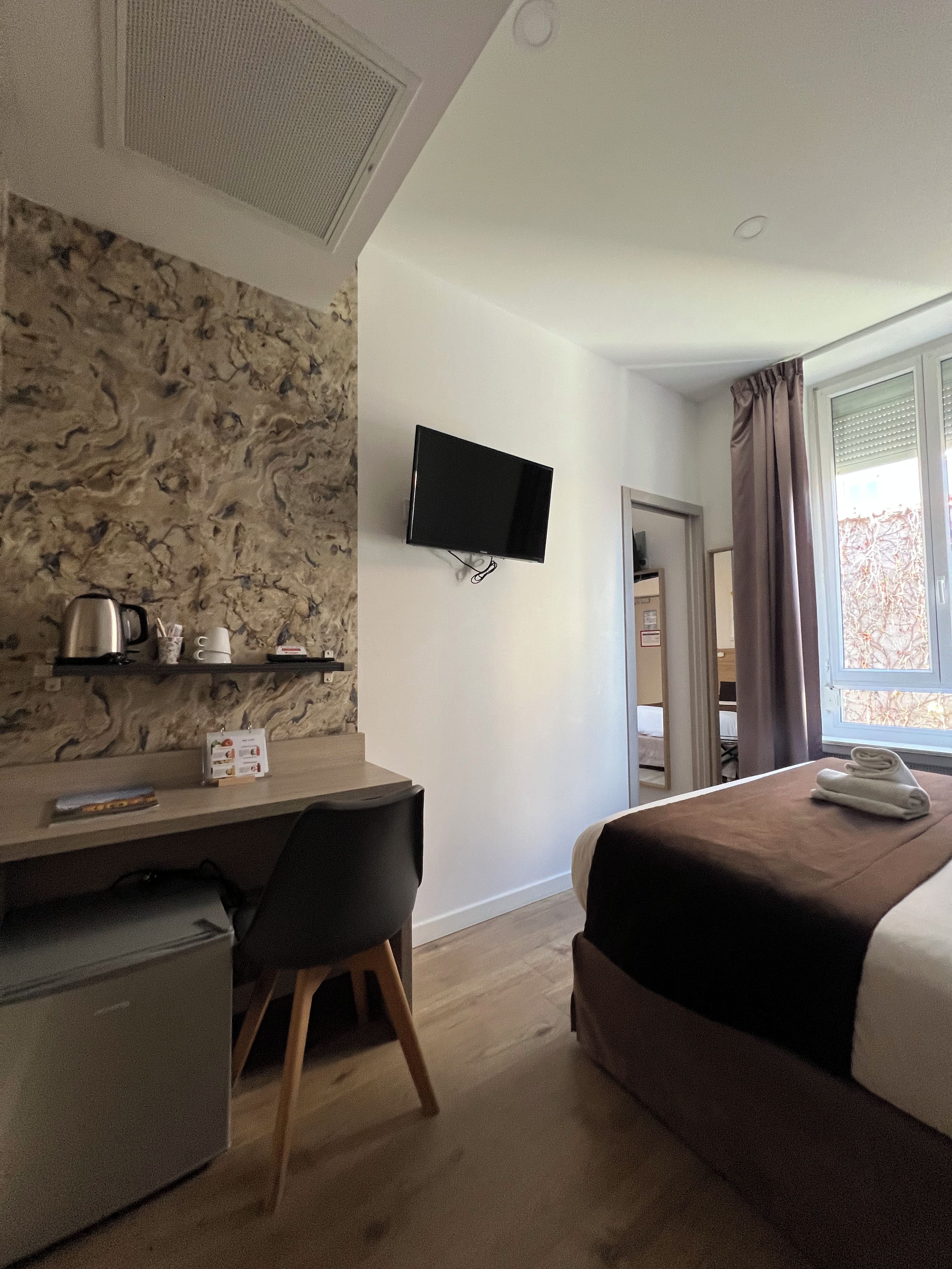 A hotel room with a flat-screen TV mounted on the wall, a desk with a chair, a bed with a brown headboard, and a window with curtains.