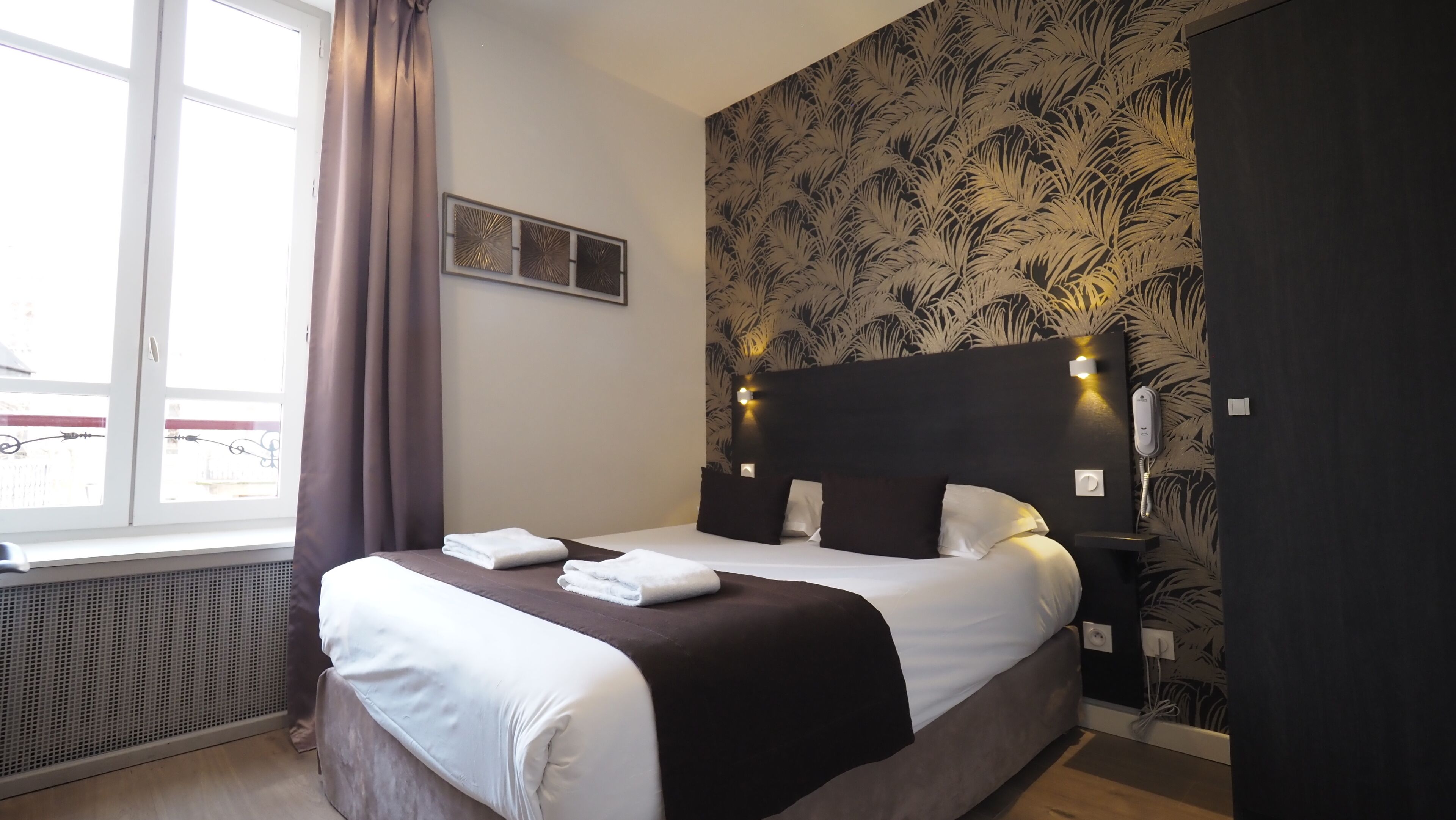 A neatly made bed with white and brown linens, a headboard with wall-mounted lights, a window with curtains, and a patterned wallpaper.