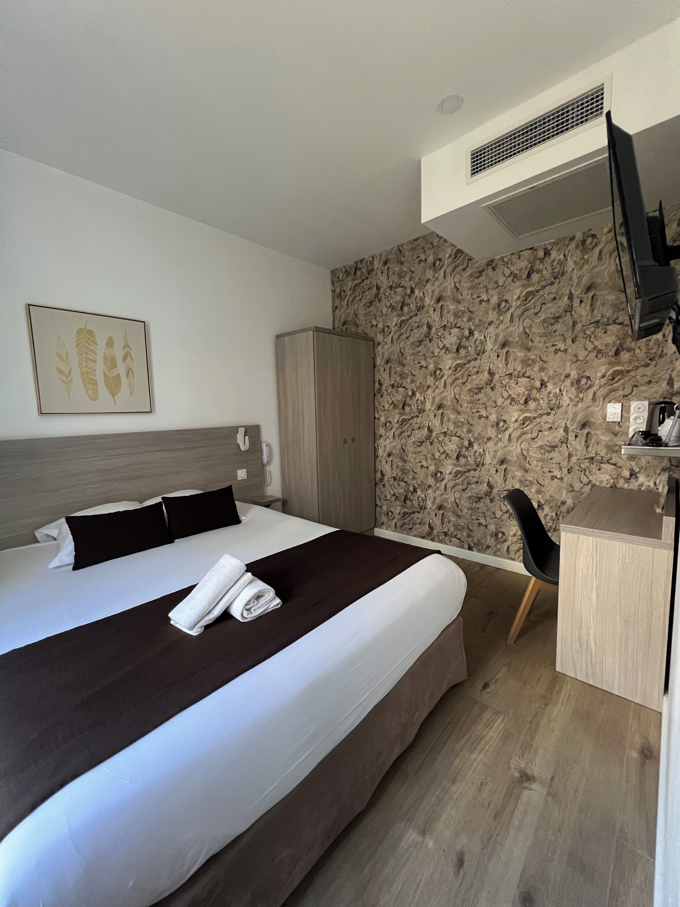 A hotel room with a bed, a desk, a wardrobe, a wall-mounted TV, and a patterned wall.