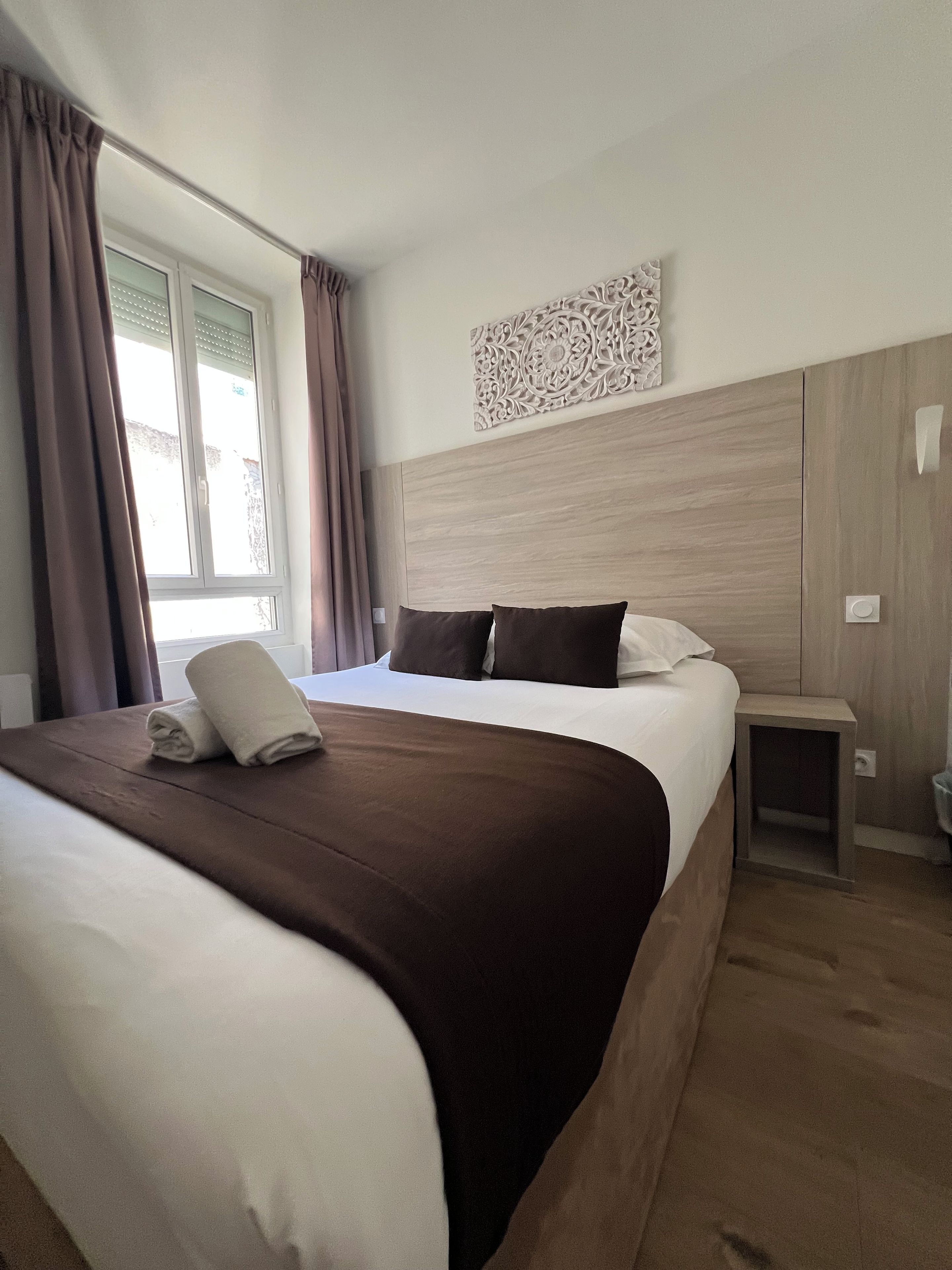 A hotel room with a large bed, a bedside table, a wall-mounted decorative piece, and a window with curtains.