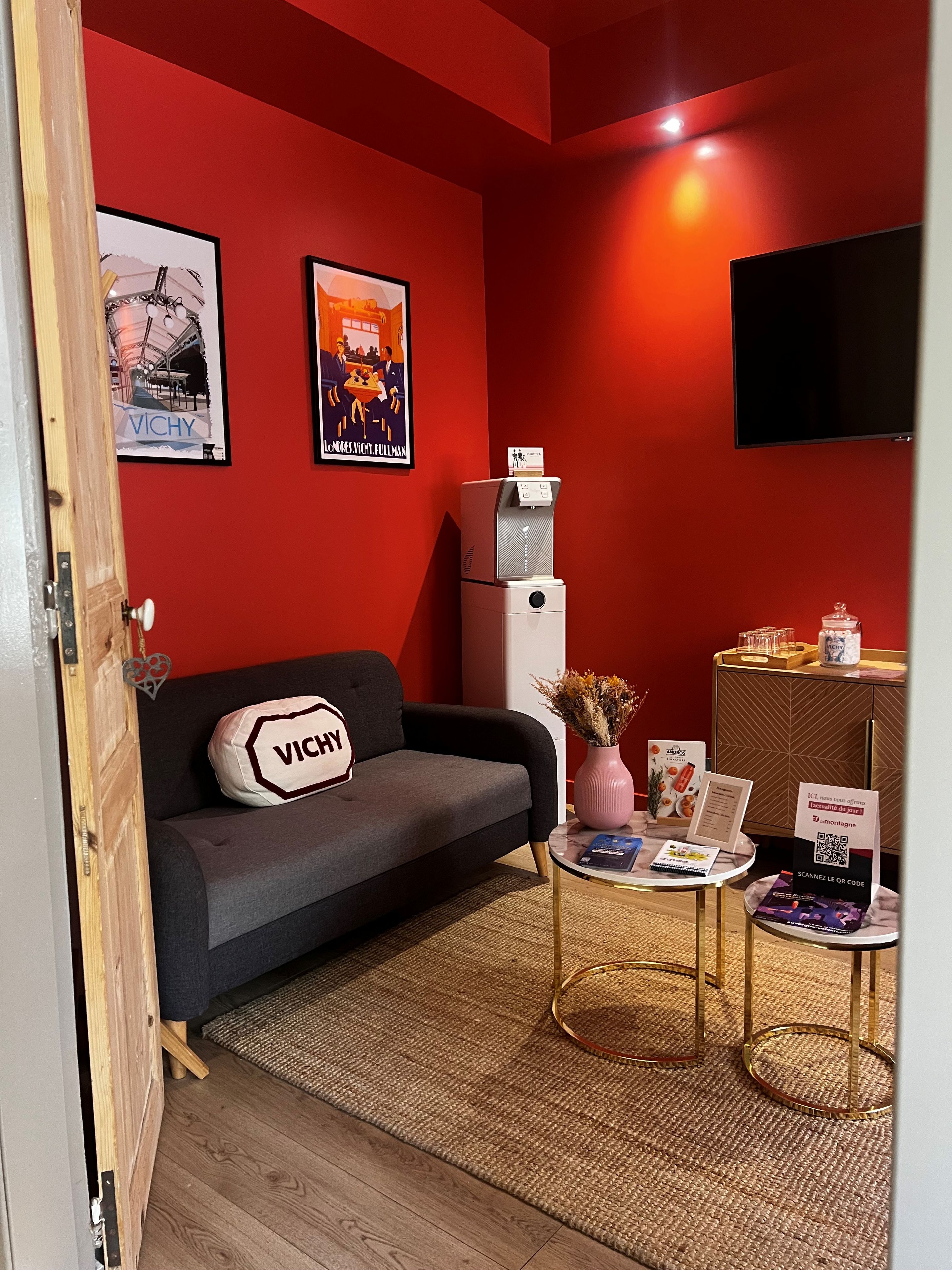A room with a red wall, a grey sofa with a 'VICHY' pillow, a small table with brochures, a vase with flowers, and a mounted television.