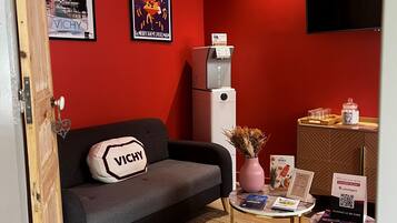 Una habitación con una pared roja, un sofá gris con un cojín 'VICHY', una mesita con folletos, un florero con flores y un televisor montado.