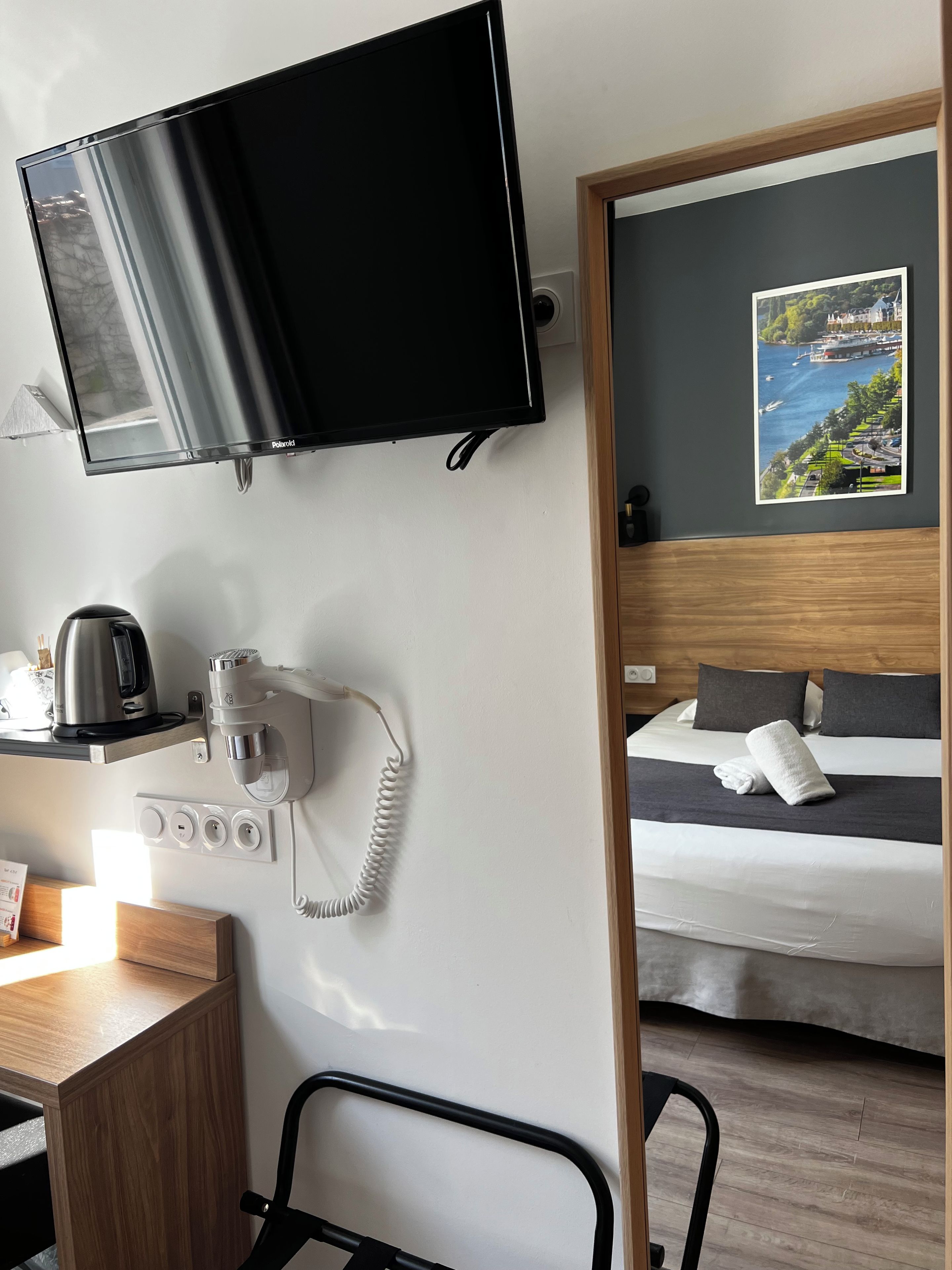 A hotel room with a bed, a flat-screen TV mounted on the wall, a coffee machine, a phone, and a painting of a landscape.