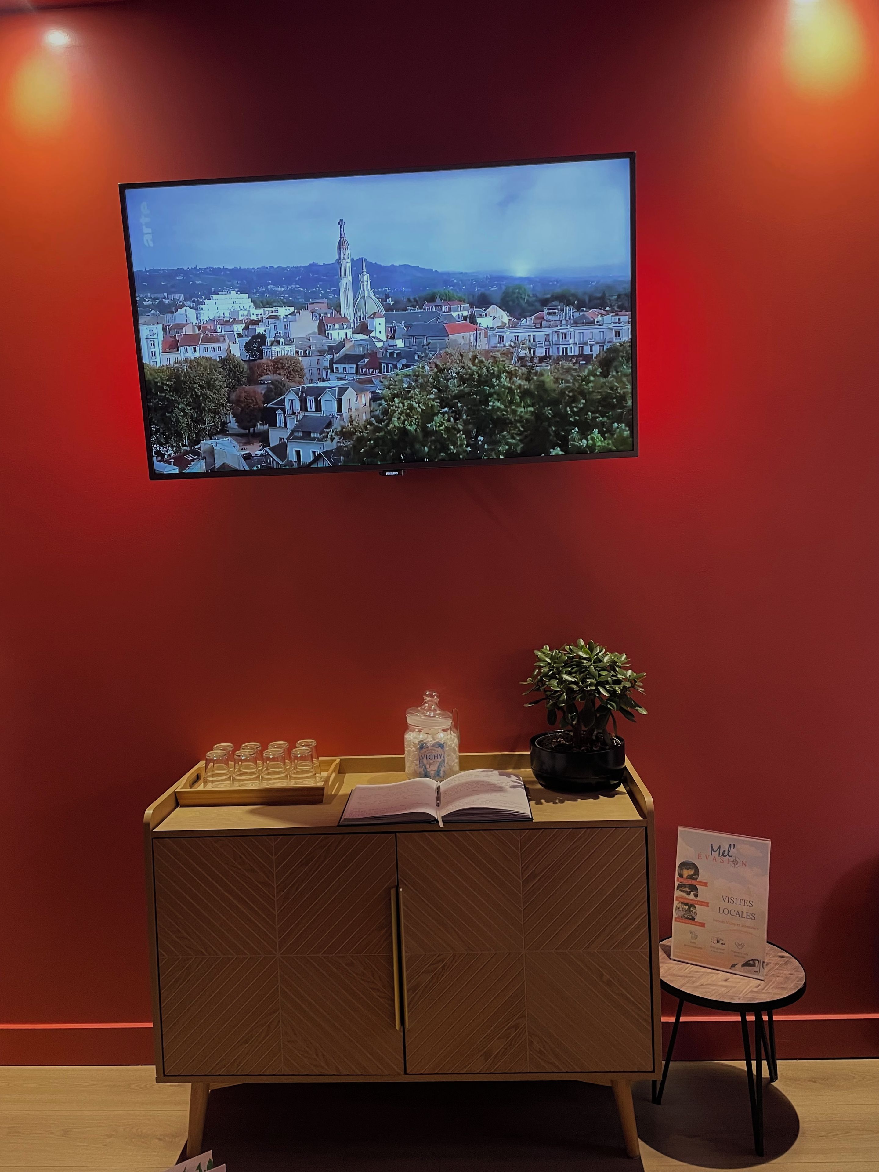 Ein Zimmer mit Fernseher, der eine Stadtansicht zeigt, einer Holzschrank mit einem Buch, einer Vase und einem kleinen Tisch mit einem Schild.