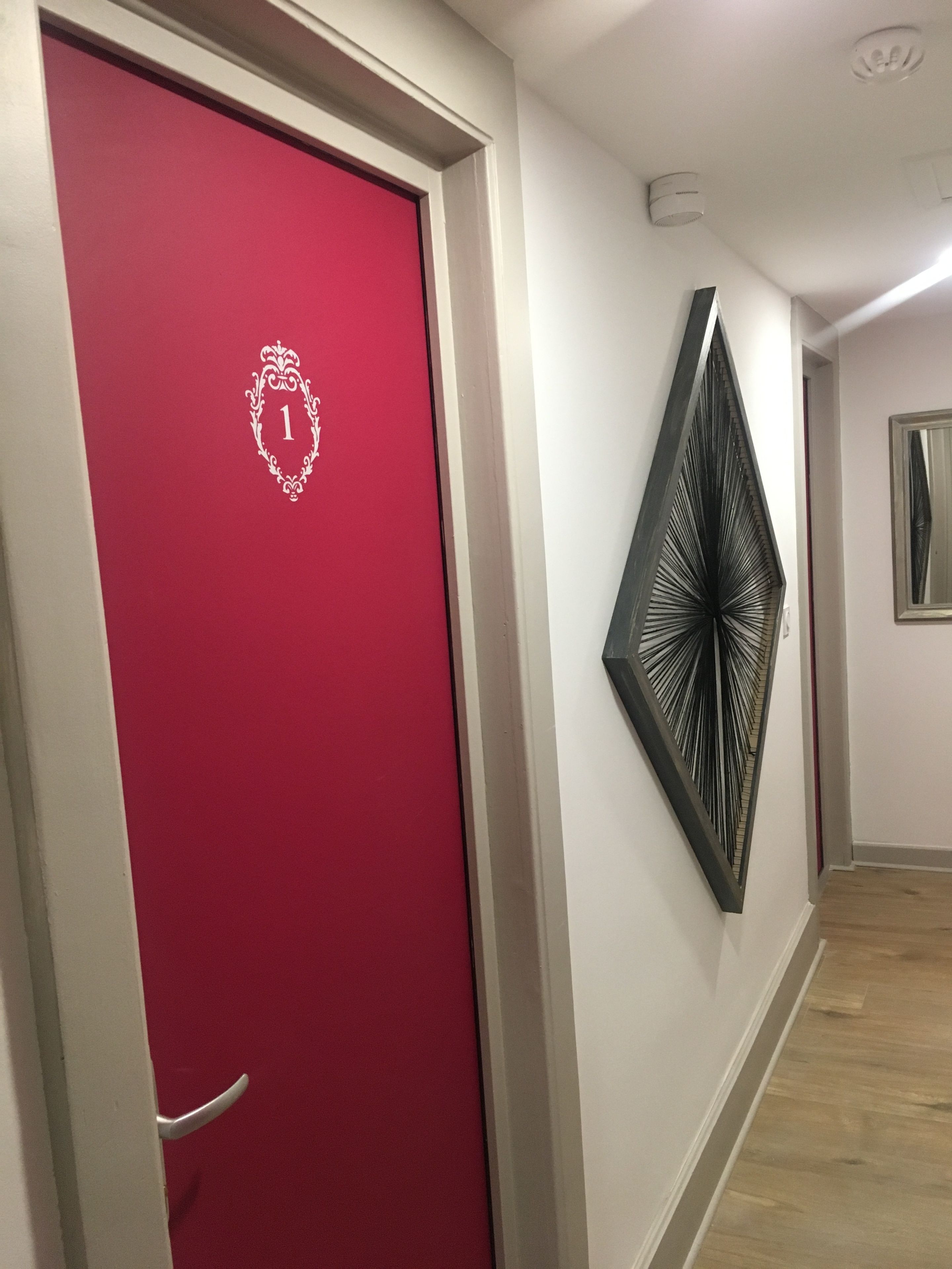 Un couloir avec une porte rouge portant un numéro, une œuvre d’art géométrique accrochée au mur et un miroir sur le mur.