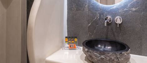 A modern bathroom with a curved shower, a round mirror, a stone sink, and a small shelf with toiletries.