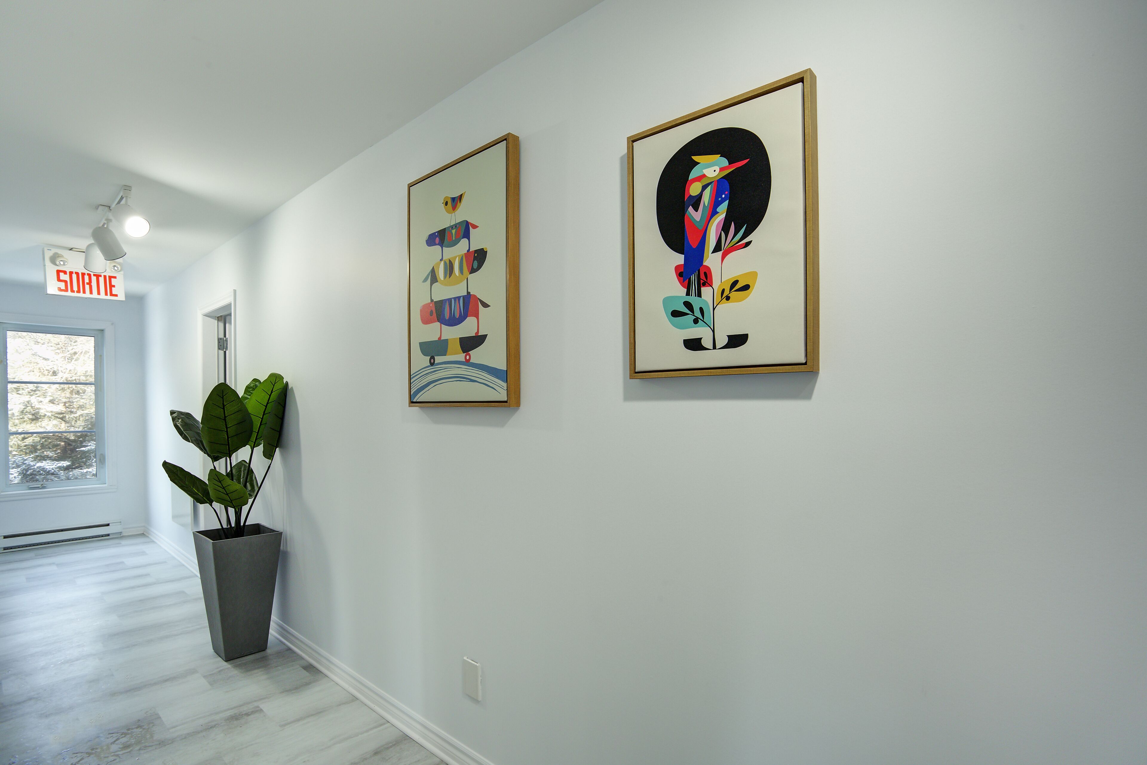 Un couloir avec deux œuvres d’art encadrées accrochées au mur, une plante en pot et une porte portant l’inscription « SORTIE ».