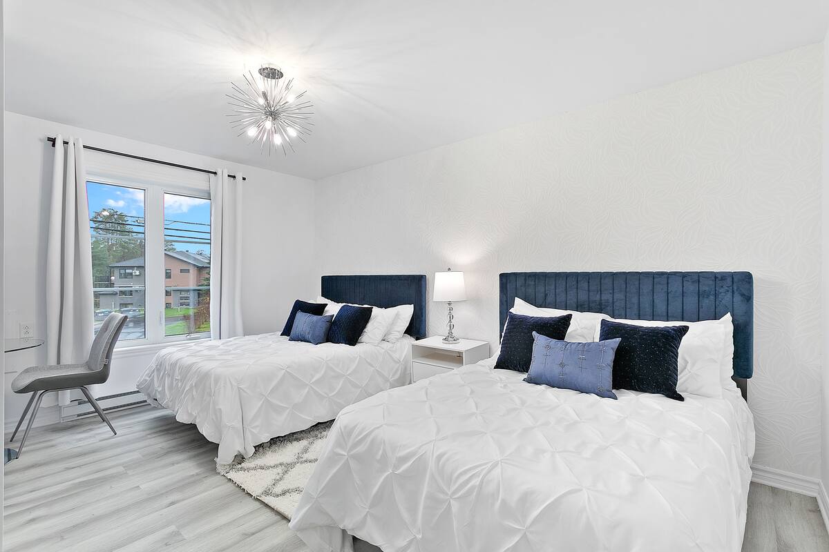 A hotel room with two beds, a chair, a nightstand, and a view of the outside.