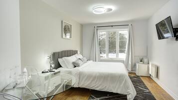 Un dormitorio moderno con cama, un escritorio de cristal, un televisor y una ventana con cortinas.