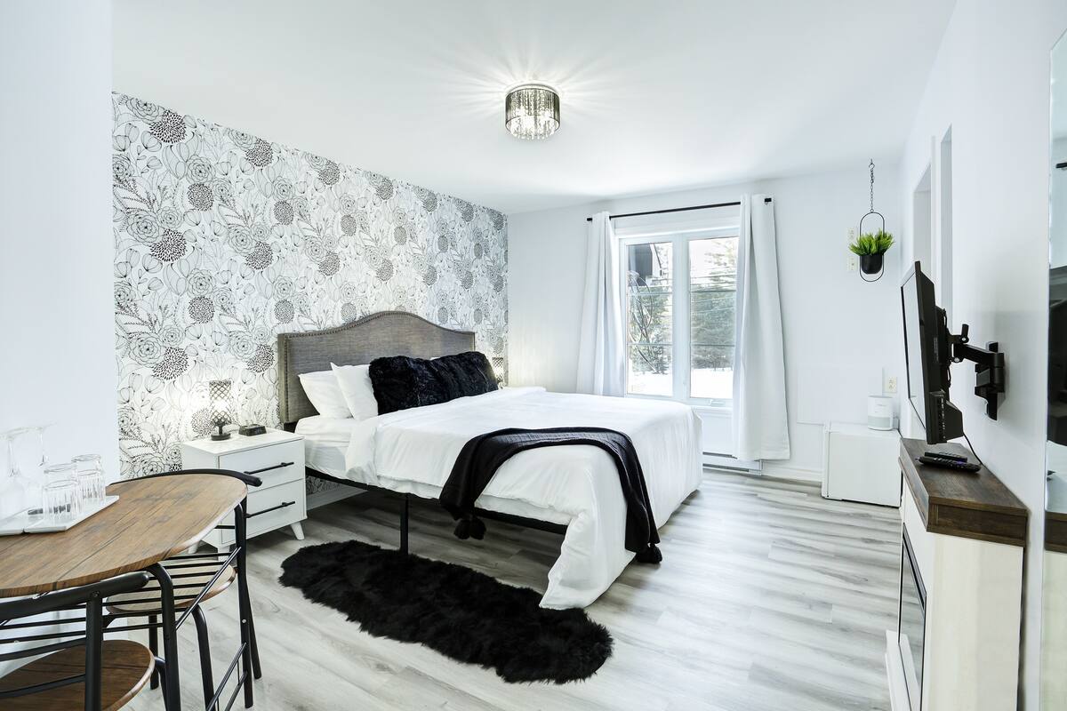 A modern bedroom with a wooden bed, white bedding, a black throw, a small table with chairs, a black furry rug, a white nightstand, a white dresser, a TV, and a window with curtains.
