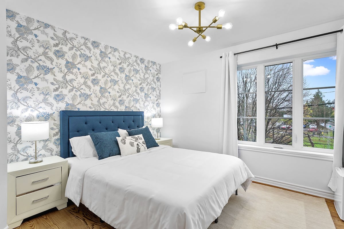 A bedroom with a large bed, a nightstand, a lamp, a chandelier, and a window with a view of trees and a road.