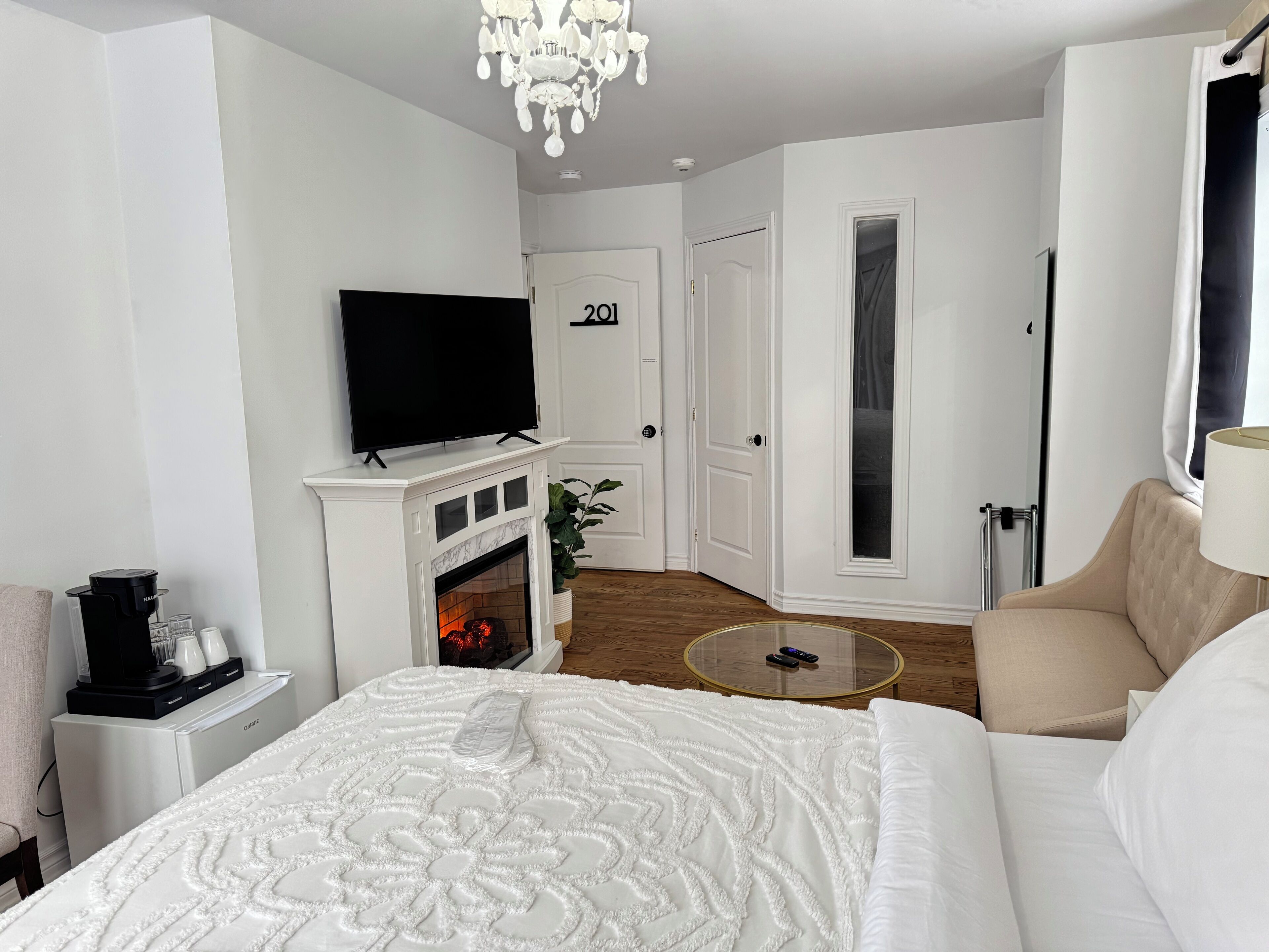 A hotel room with a bed, a TV mounted above a fireplace, a door labeled "201", and a round glass table.
