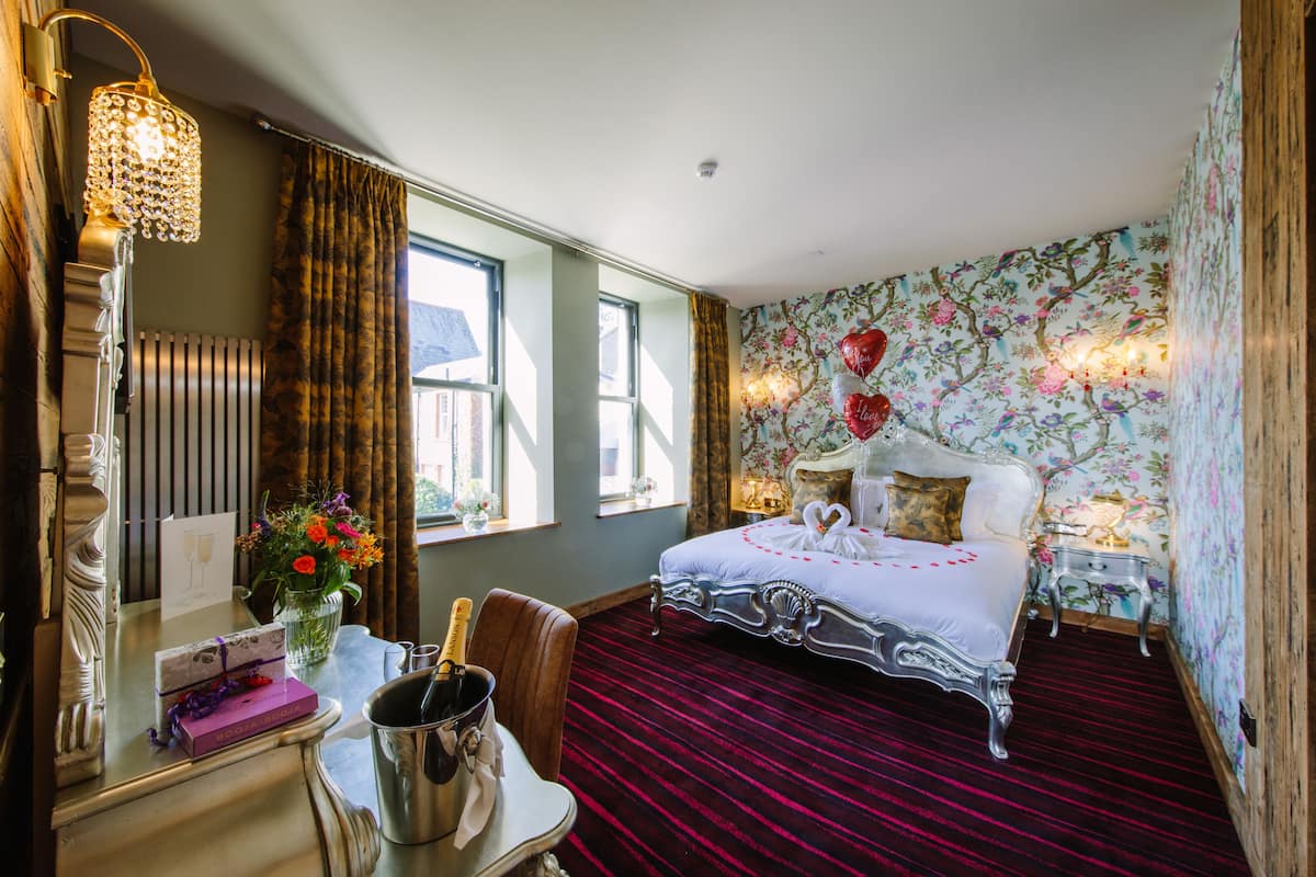 Une chambre d’hôtel avec un grand lit, une coiffeuse avec un miroir, une table de chevet ornée d’un vase de fleurs et un sol recouvert d’une moquette rouge.