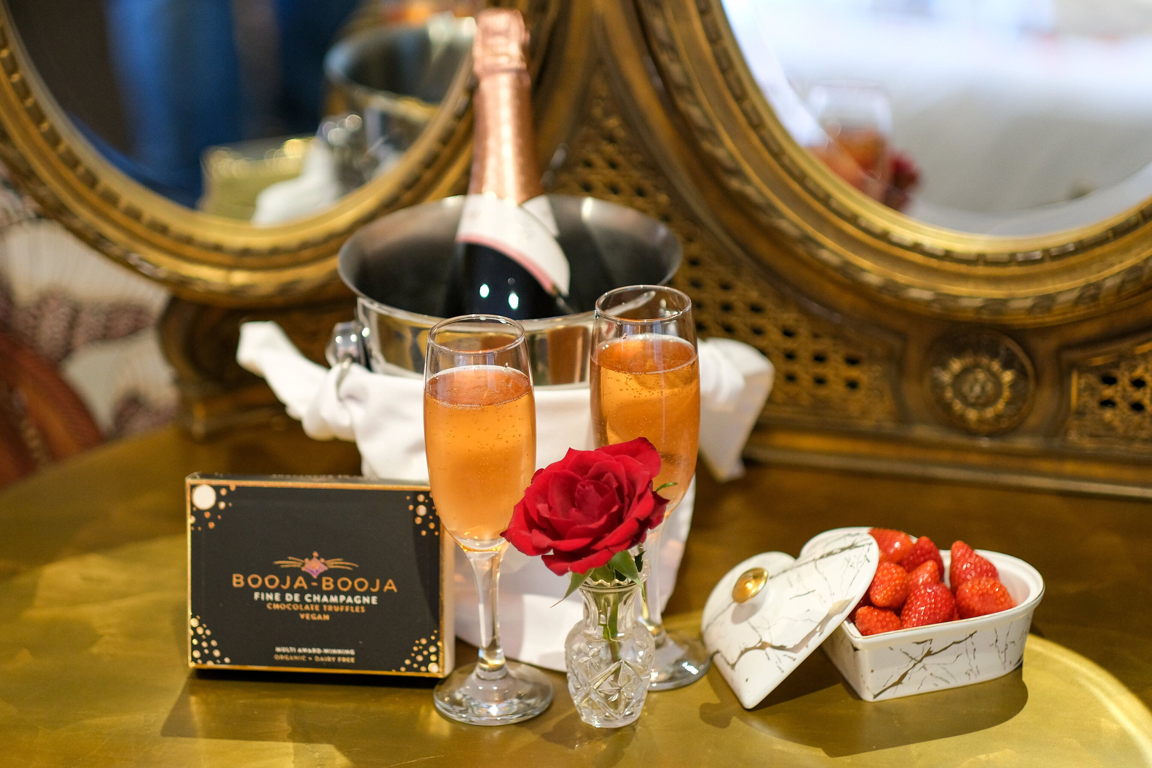 A table with a bottle of champagne, two glasses of champagne, a red rose, a box of chocolate truffles, and a bowl of strawberries.