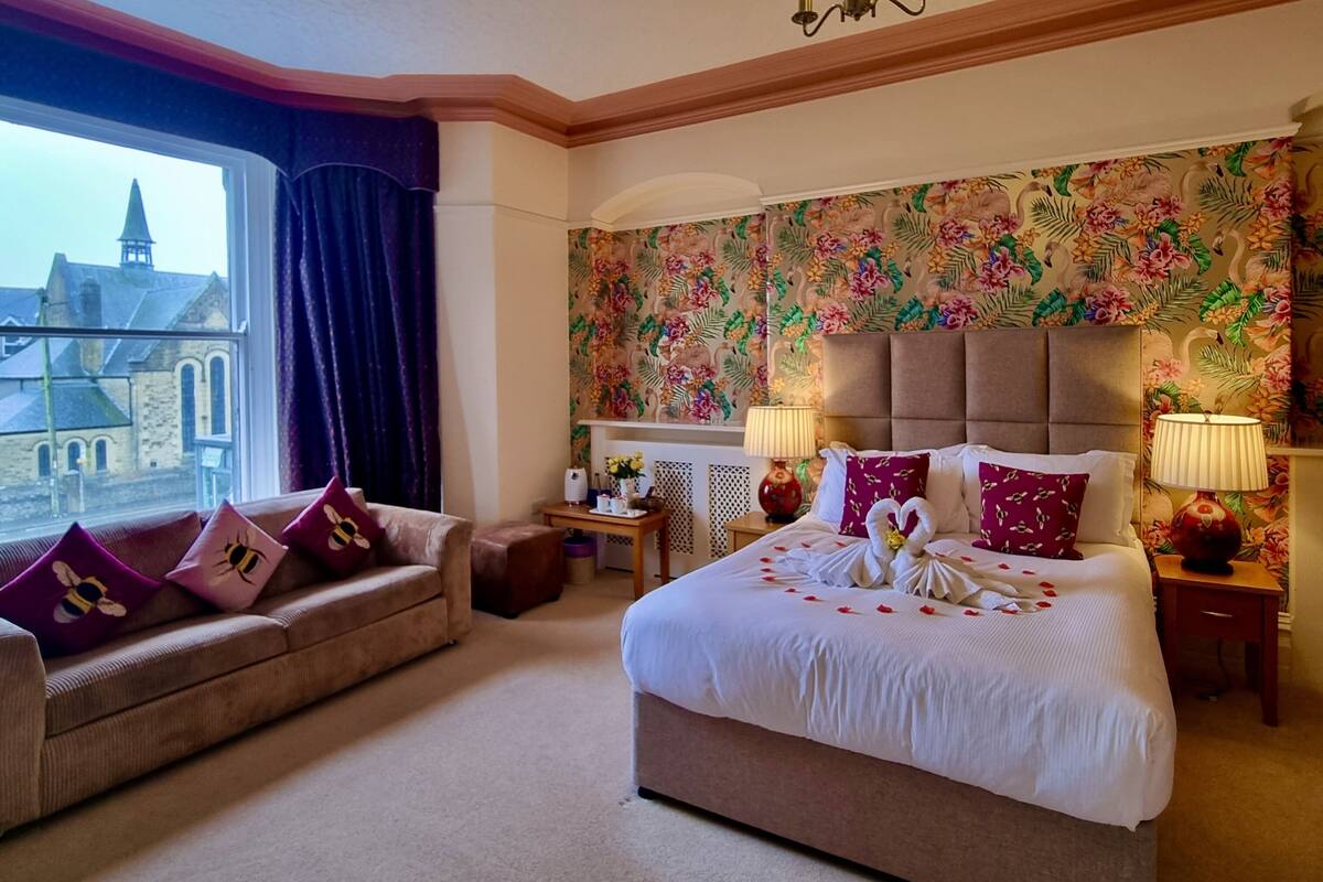 Une chambre d’hôtel avec un grand lit, un canapé, une fenêtre donnant sur un immeuble, un papier peint à motifs floraux et deux lampes de chevet.