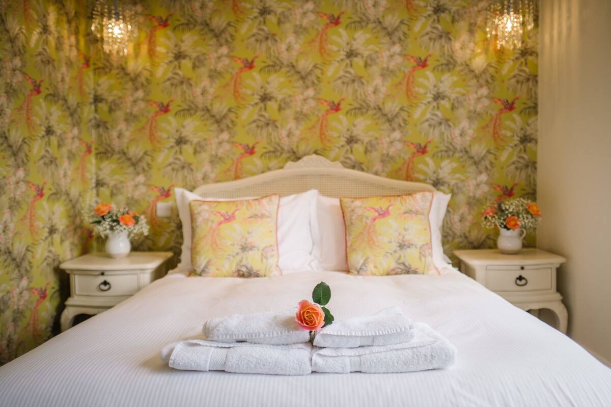 Un lit bien fait, avec du linge de lit blanc et une rose posée sur les oreillers, le tout mis en valeur par un papier peint à motifs floraux.