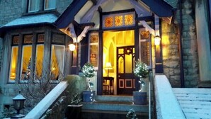 A stone building with a steeply pitched roof, a wooden front porch with a staircase, and a lit interior visible through the windows. - Lymestone Boutique Accommodation (Grange-over-Sands)