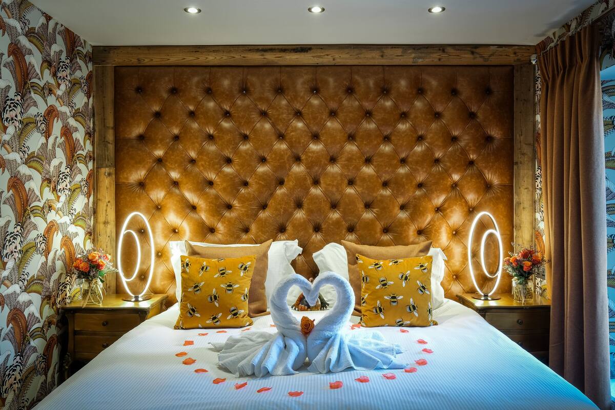 Une chambre d’hôtel avec une tête de lit capitonnée, un papier peint à motifs floraux, un lit agrémenté d’une décoration en forme de cœur faite avec des serviettes, et une table de chevet avec un miroir.