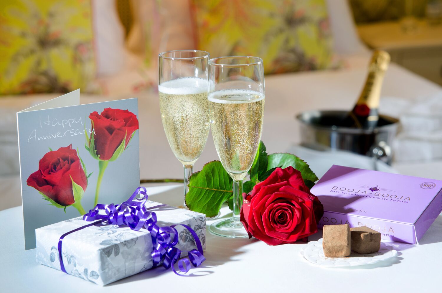 Una mesa con una tarjeta de felicitación, dos copas de champán, una rosa roja, una caja de trufas de chocolate y un regalo envuelto en papel floral.