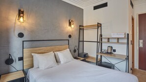 A modern hotel room with a large bed, two bedside tables, a wall-mounted shelf with towels, and a small desk with a kettle and cups. - Hotel Auguste (Paris)