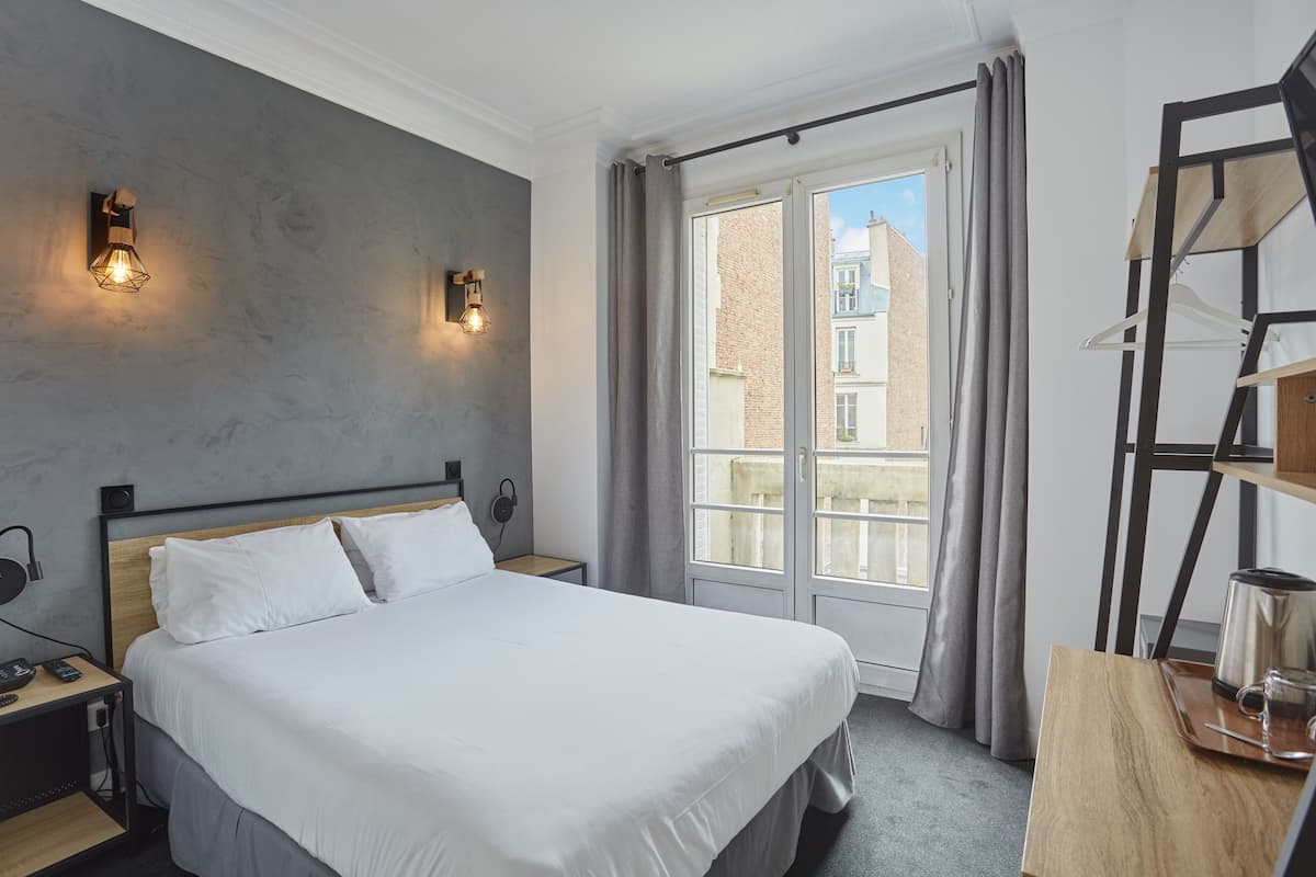 A modern hotel room with a large bed, two bedside tables, a desk with a kettle, and a view of the city through a large window.