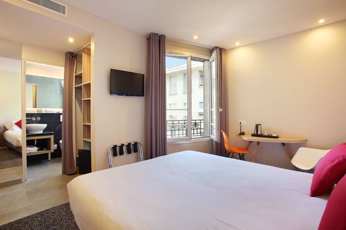 A hotel room with a large bed, a desk with a chair, a TV mounted on the wall, and a balcony with a view.