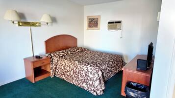 A hotel room with a bed, bedside table, lamp, air conditioning unit, and a desk with a computer.
