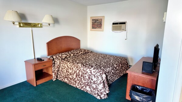A hotel room with a bed, bedside table, lamp, air conditioning unit, and a desk with a computer.