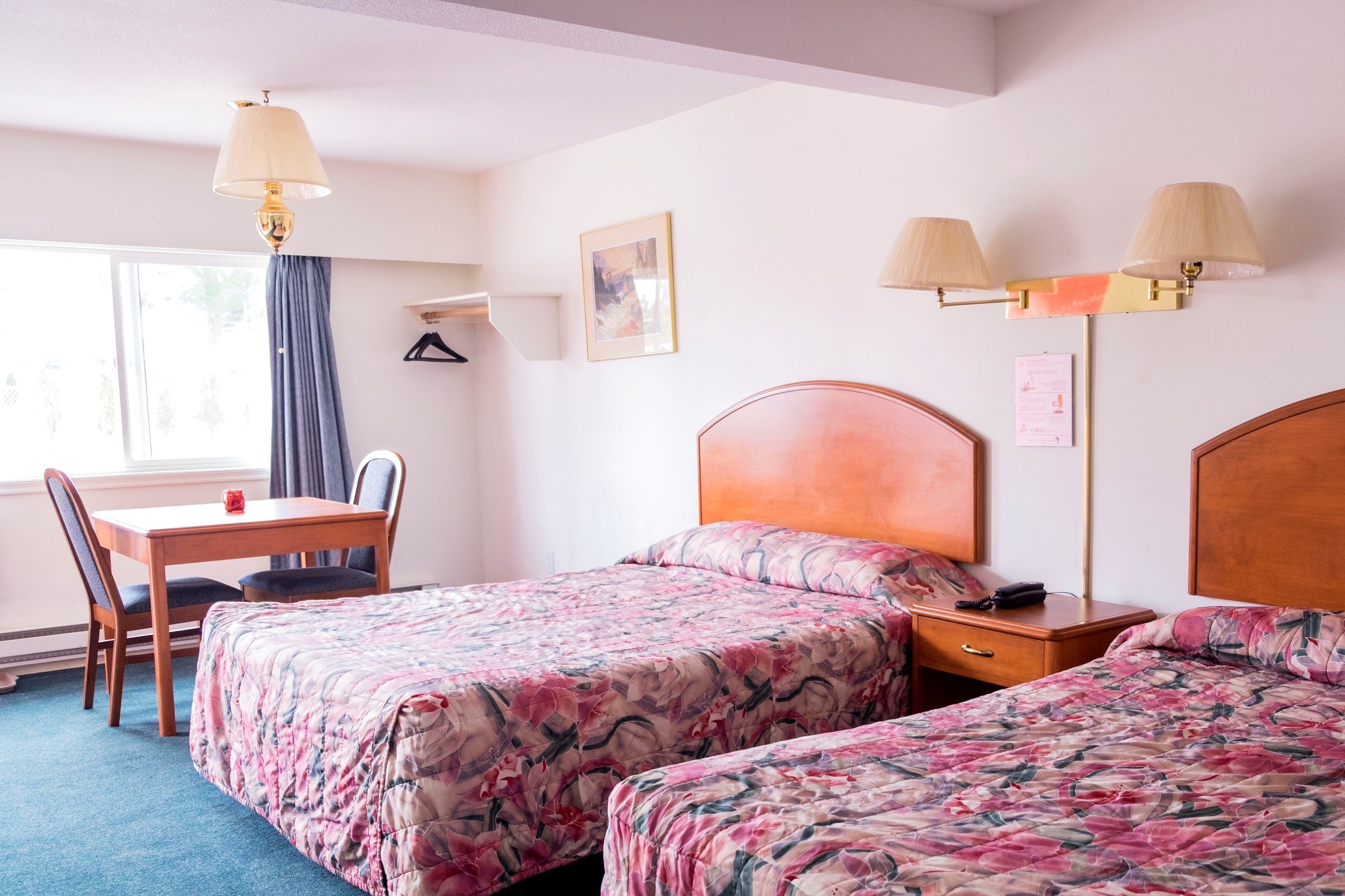 Une chambre d’hôtel avec deux lits, un bureau et une chaise. Il y a deux lampes fixées au mur et une fenêtre avec des rideaux.
