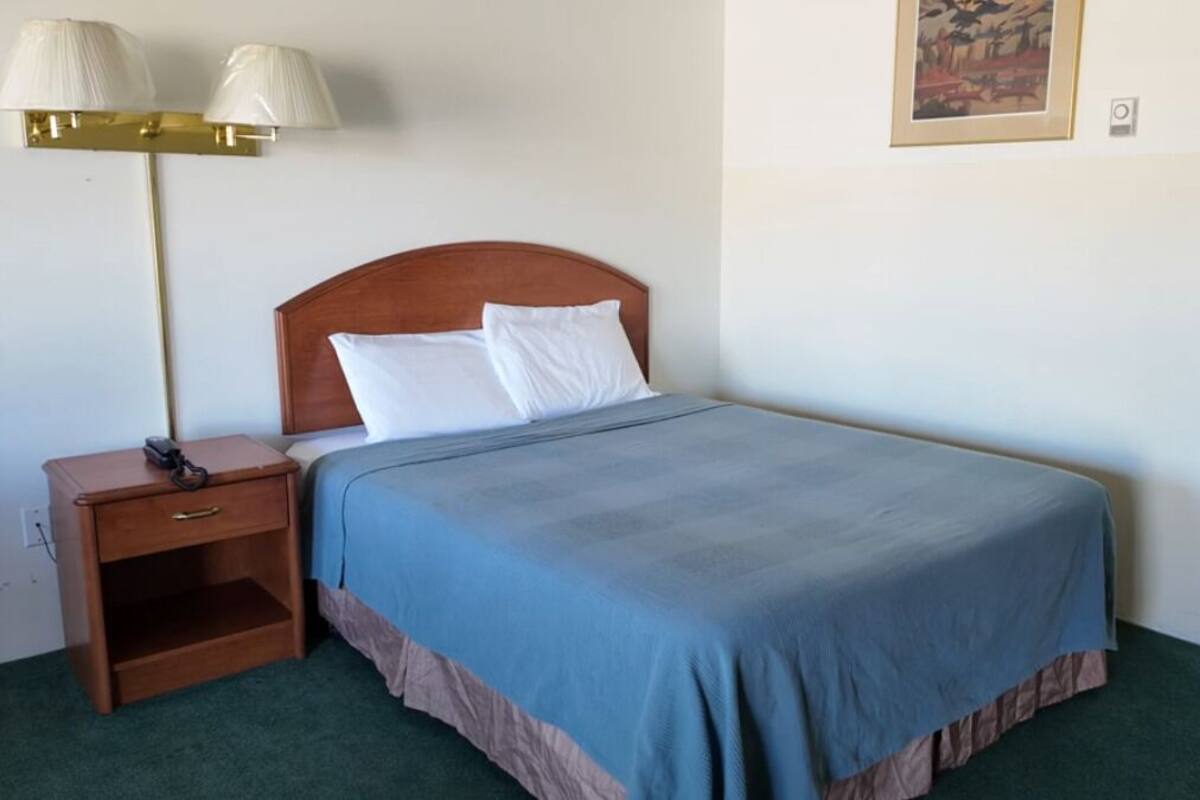 A neatly made bed with a blue bedspread, a wooden nightstand with a lamp, and a framed picture on the wall.