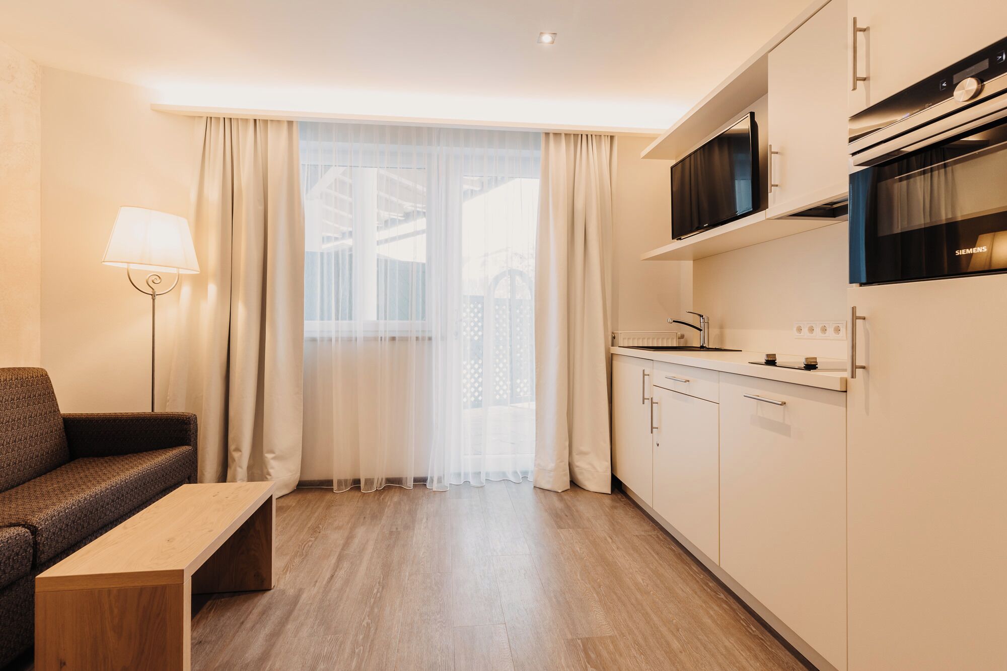 A modern hotel room with a kitchenette, a sofa, a small wooden table, and a large window with sheer curtains.