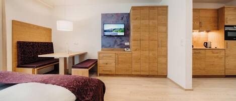 Habitación de hotel pequeña con cama, escritorio, televisor, kitchenette y armarios de madera.