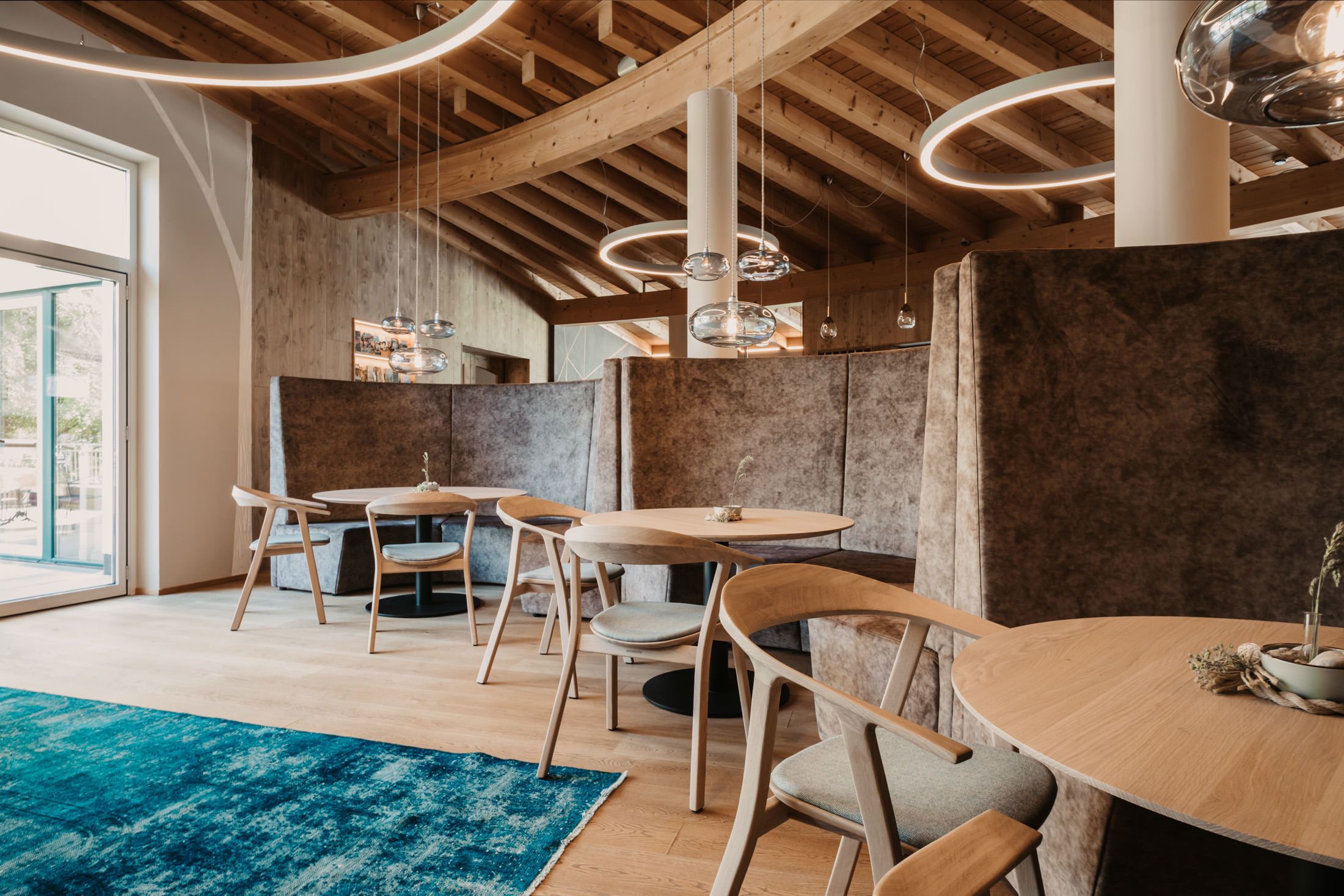 A modern dining area with wooden tables and chairs, a blue rug, and a high ceiling with exposed beams and pendant lighting.